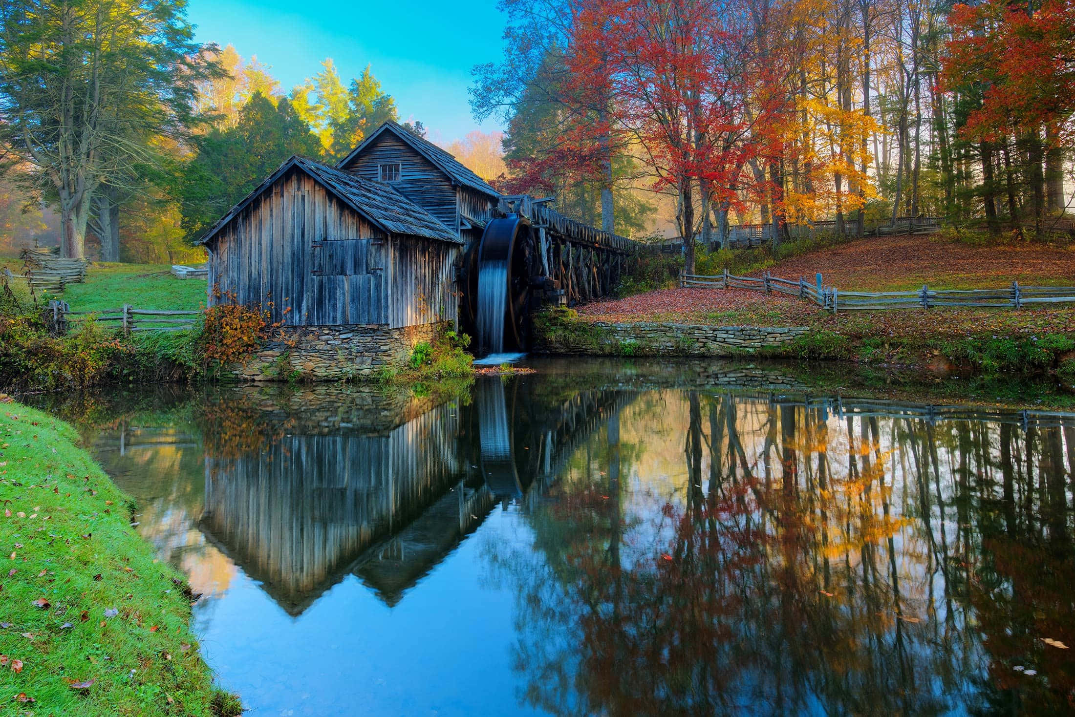Blue Ridge Parkway 桌布 壁紙 — 一座質樸的木製水車磨坊，帶有流動的水車，完美地倒映在平靜的池塘中，周圍環繞著充滿活力的秋葉。
