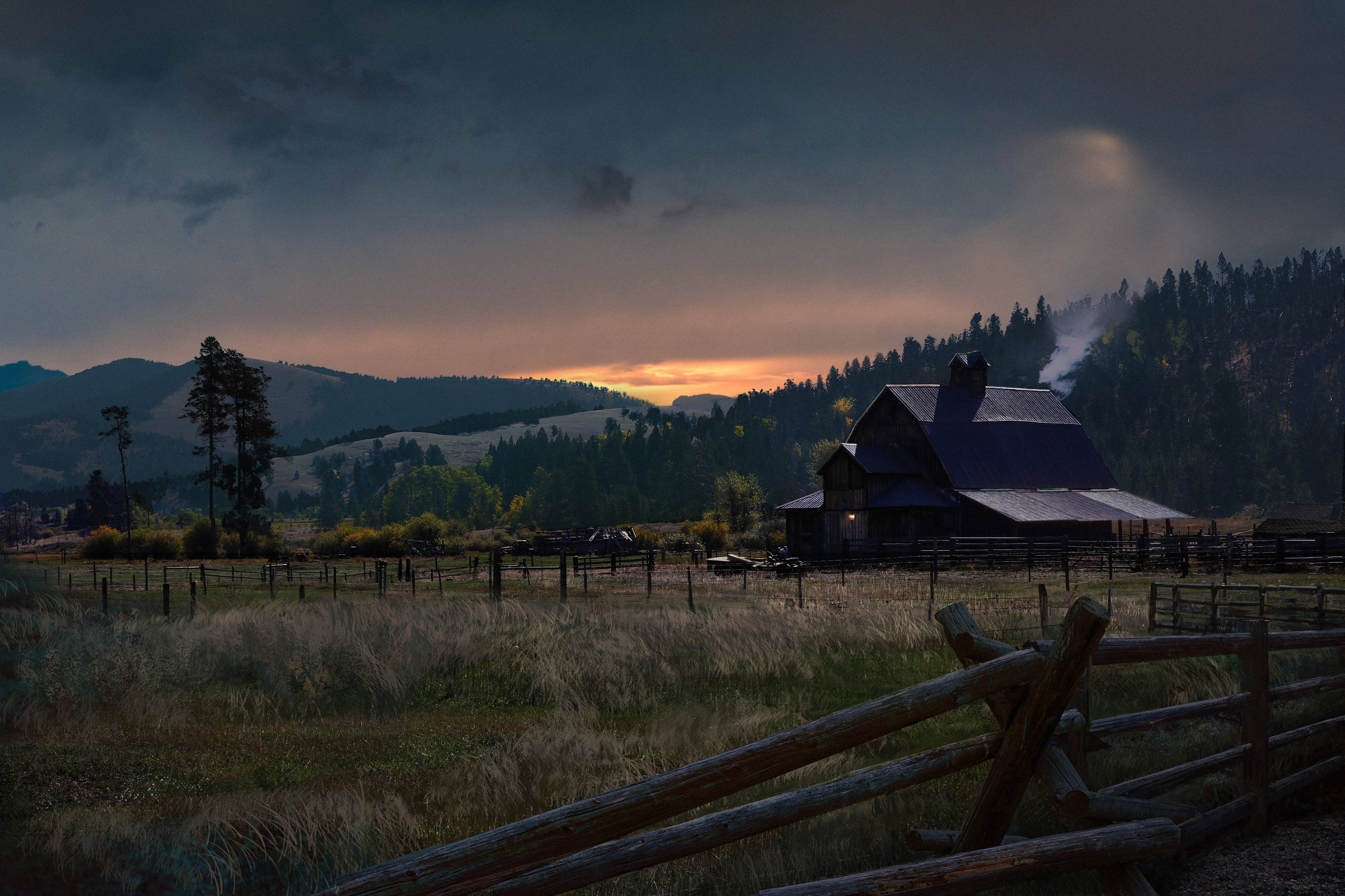 Far Cry 5 桌布 壁紙 — 一座質樸的木製穀倉在暮色天空中冒出煙霧，天空有橙色雲彩，背景是遠山和松林。