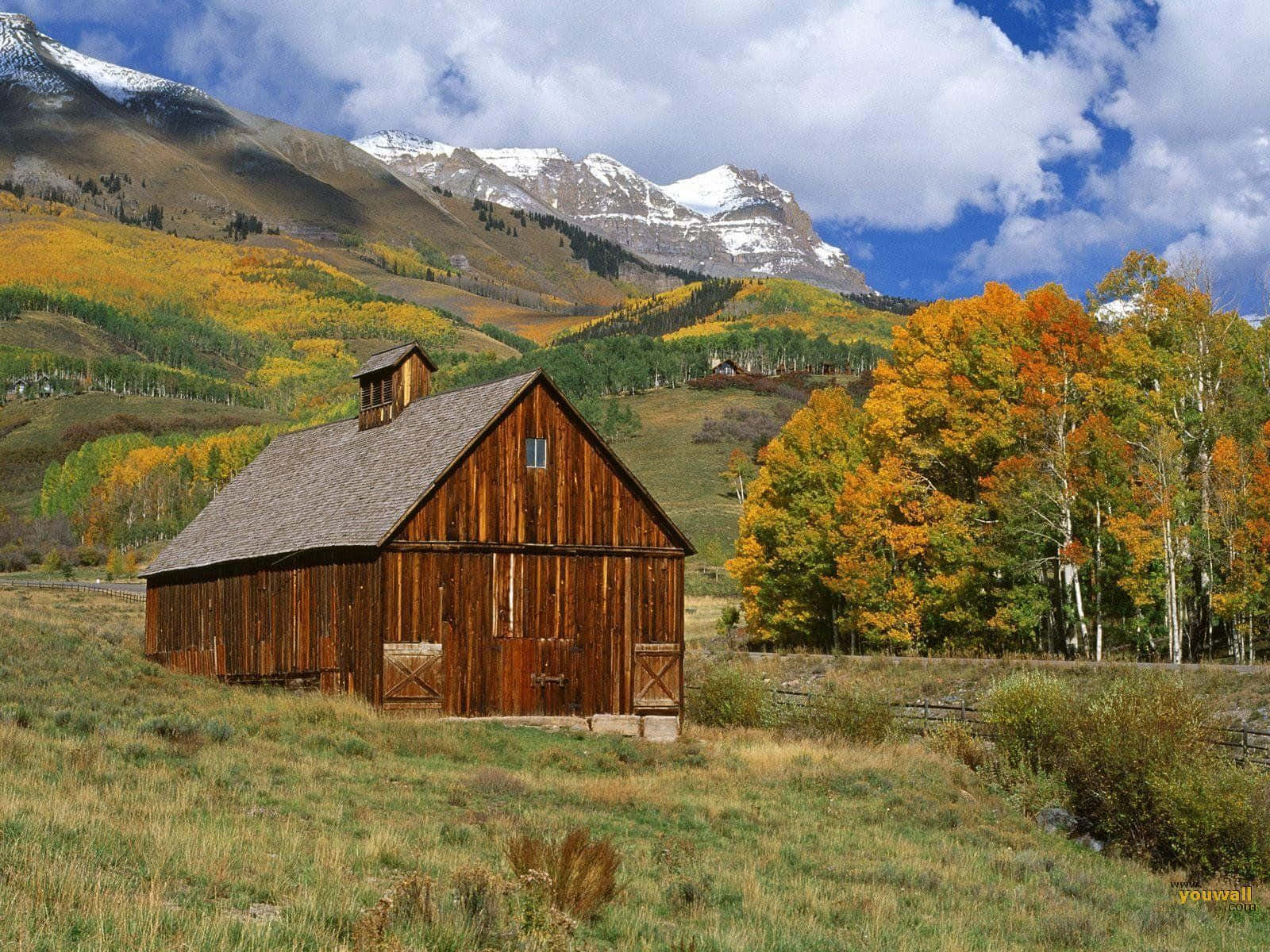 Fall Barn 桌布 壁紙 — 一座質樸的木製穀倉矗立在長滿青草的田野中，周圍環繞著秋樹和積雪覆蓋的山脈，天空部分多雲。