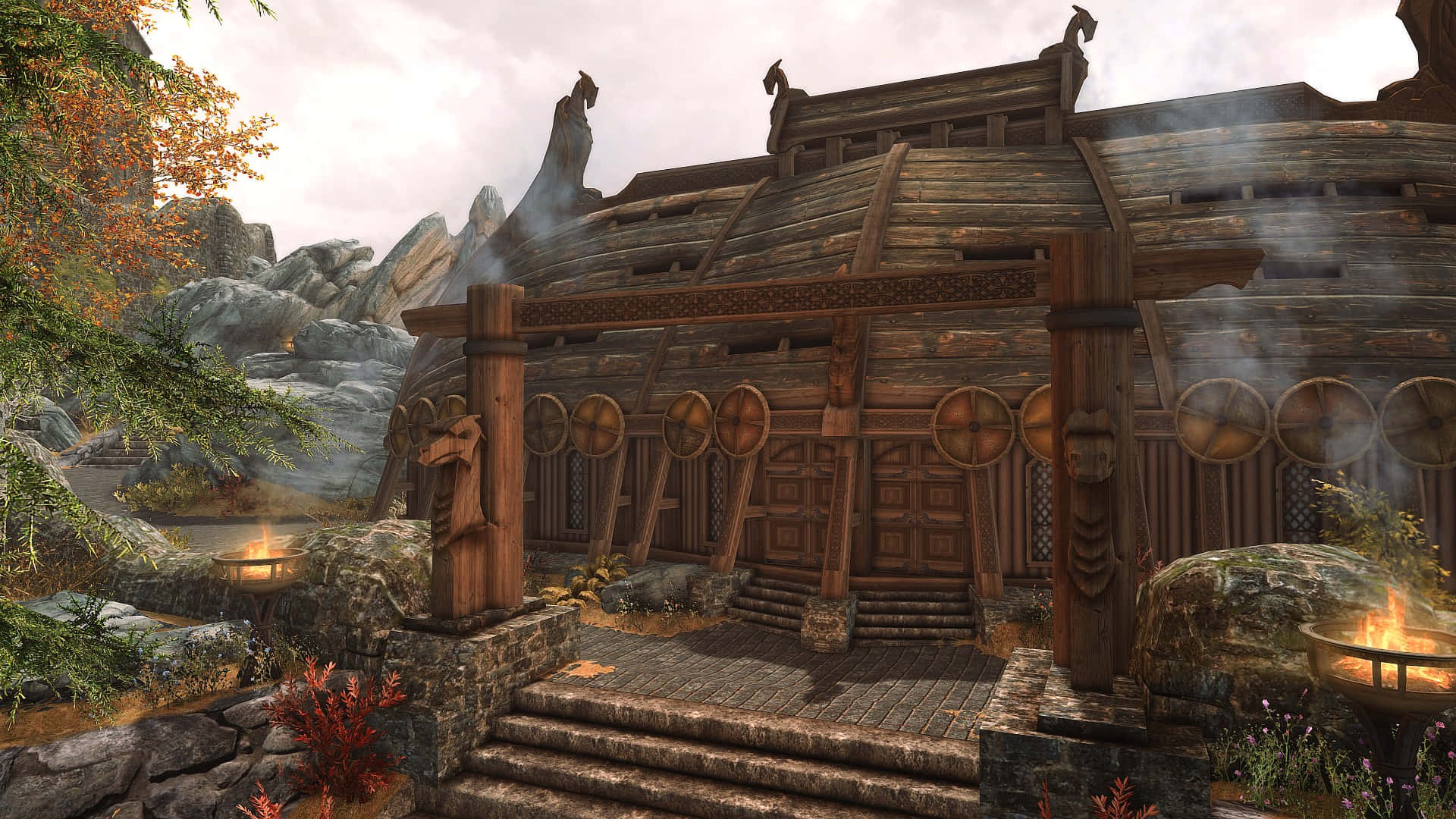 Skyrim Whiterun 桌布 壁紙 — 一座質樸的木製維京長屋，屋頂升起裊裊炊煙，坐落在秋日樹木和岩石地形之間。