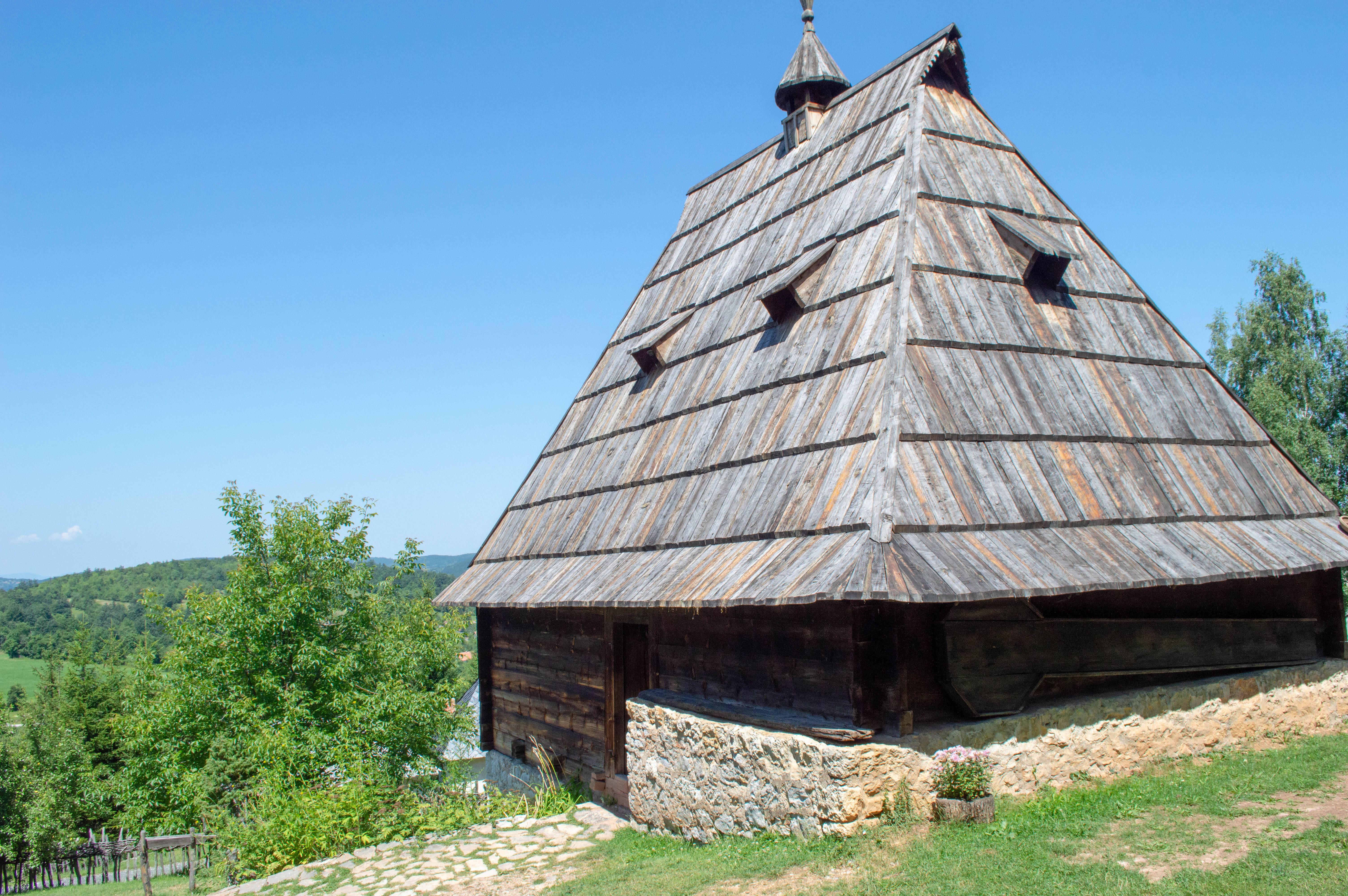 Serbia 桌布 壁紙 — 一座質樸的木造教堂，帶有陡峭的木瓦屋頂，矗立在晴朗藍天下的草地山坡上。