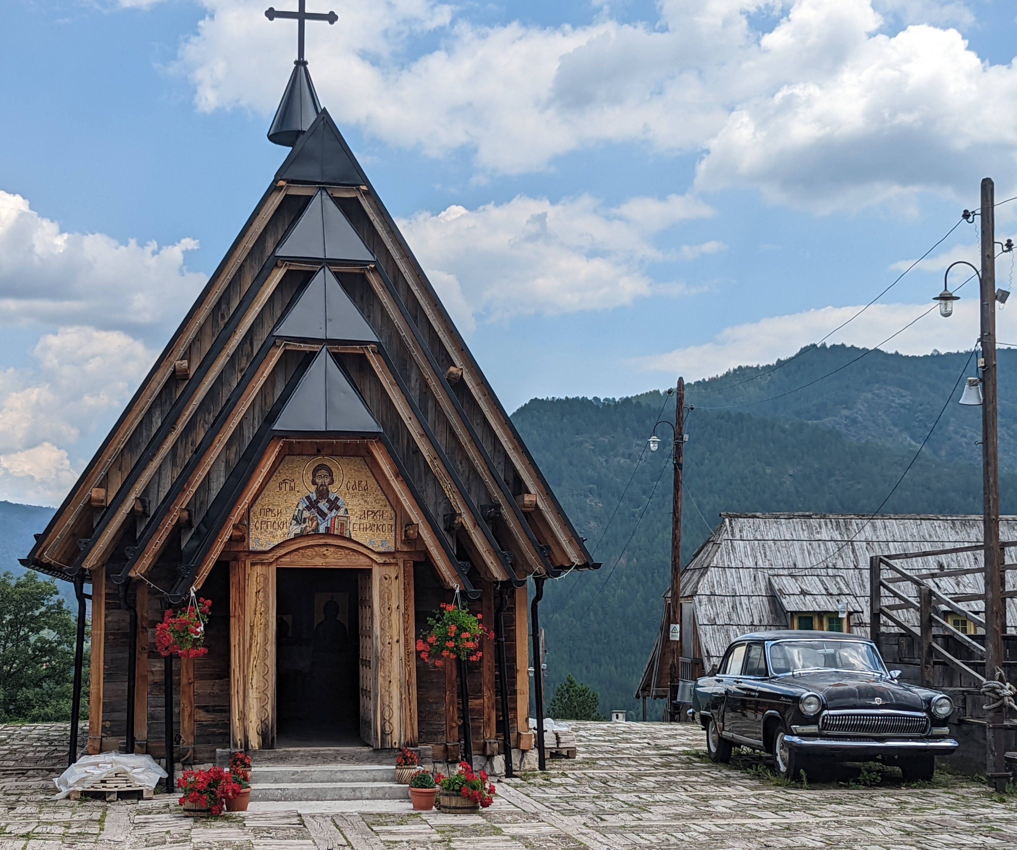 Serbia 桌布 壁紙 — 一座質樸的木造教堂，擁有陡峭的屋頂和十字架，矗立在石頭廣場上，旁邊有一輛復古汽車，天空部分多雲。