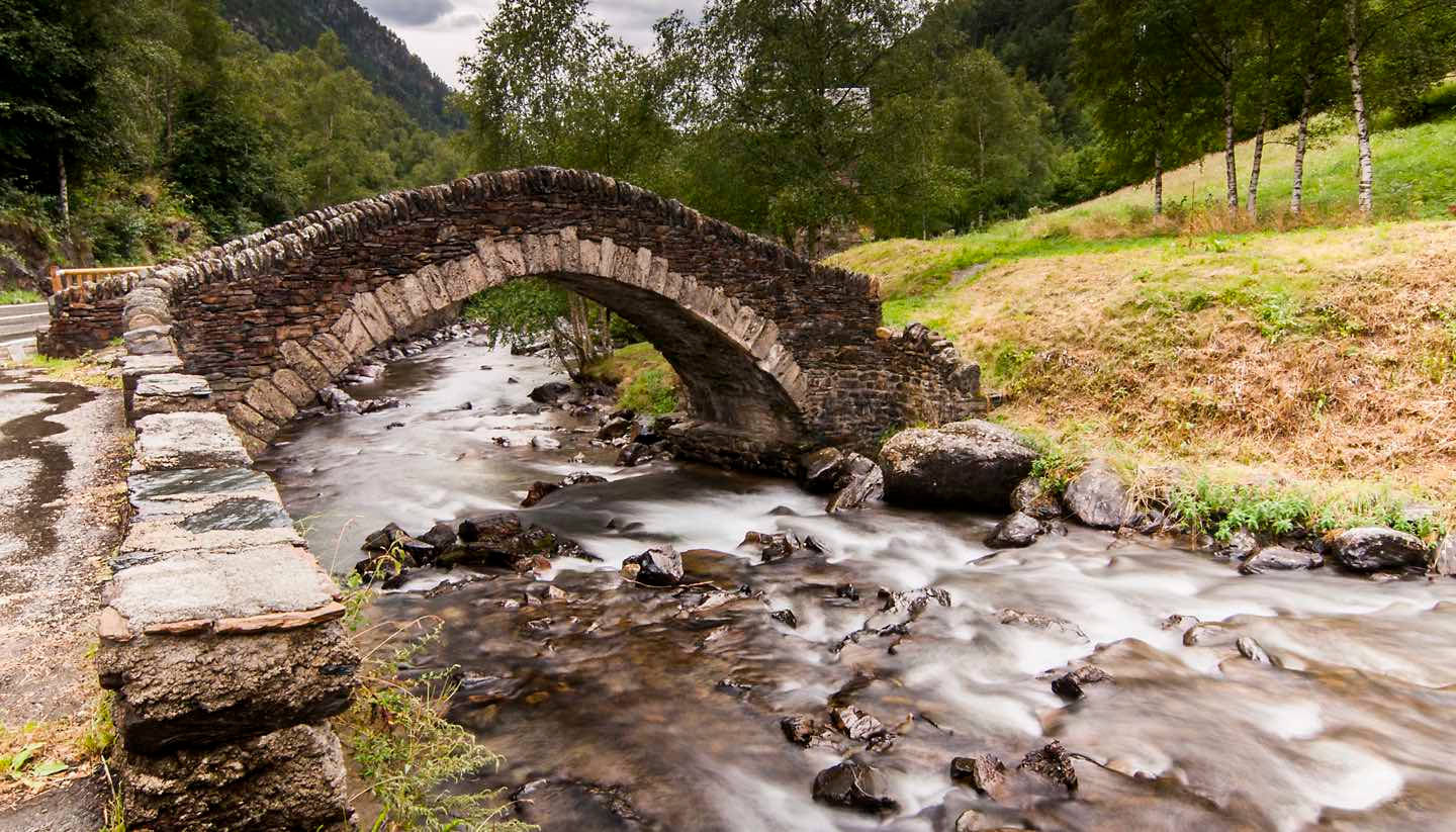 bridge 桌布 壁紙 — 一座質樸的石拱橋橫跨湍急的山澗，周圍是茂密的綠色森林和草坡。