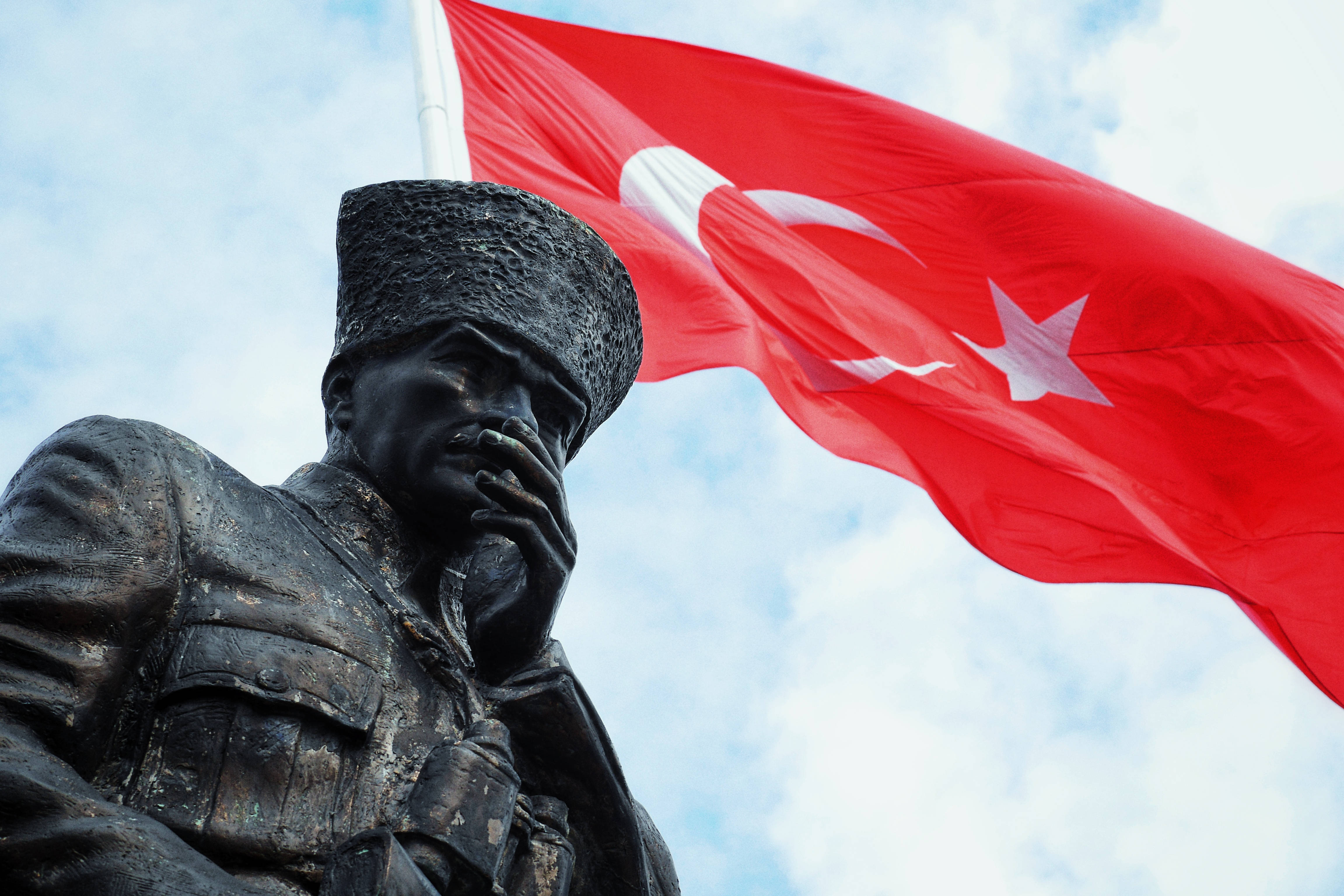 Ataturk 桌布 壁紙 — 一座身穿制服、手掩嘴巴的士兵青銅雕像，矗立在飄揚的紅色土耳其國旗下，國旗上有白色新月和星星，背景是陰雲密布的天空。