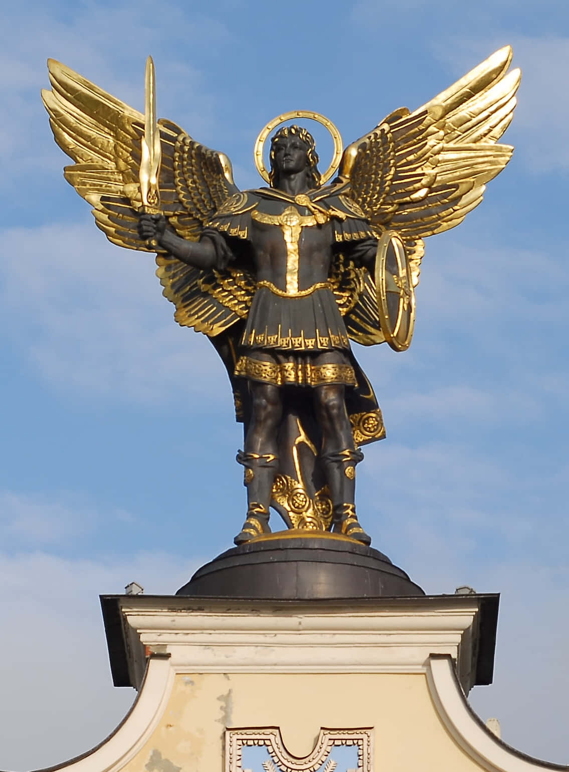 Saint Michael 桌布 壁紙 — 一座身穿盔甲、擁有金色翅膀的雄偉天使雕像，在清澈的藍天下手持劍與盾。