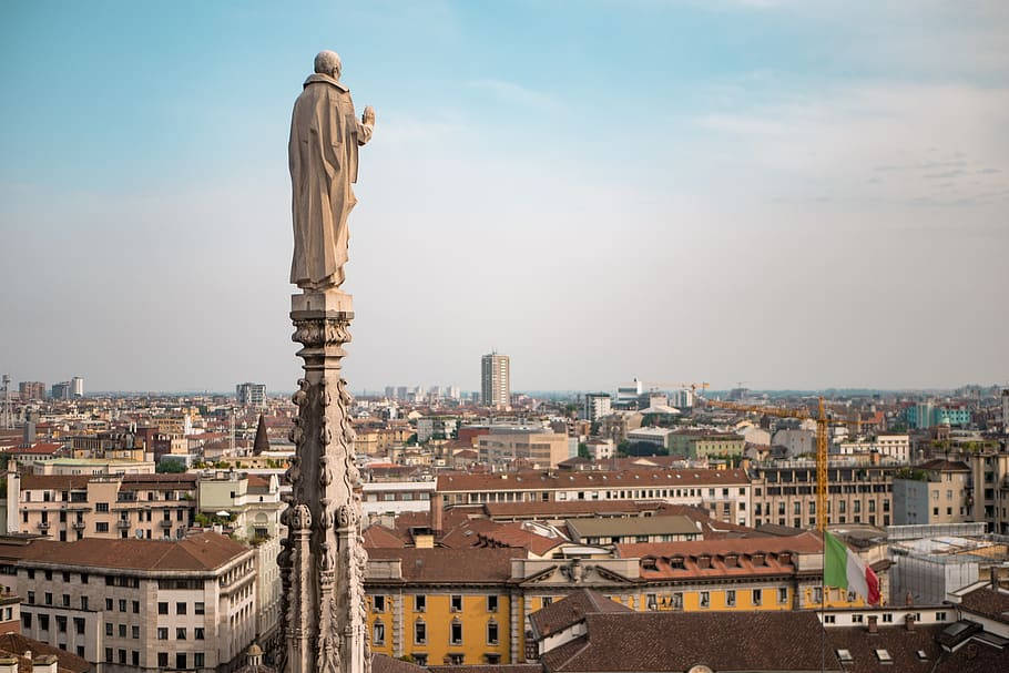 Milan 桌布 壁紙 — 一座身穿長袍的人物石像矗立在尖塔頂端，俯瞰著朦朧藍天下廣闊的歐洲城市景觀。
