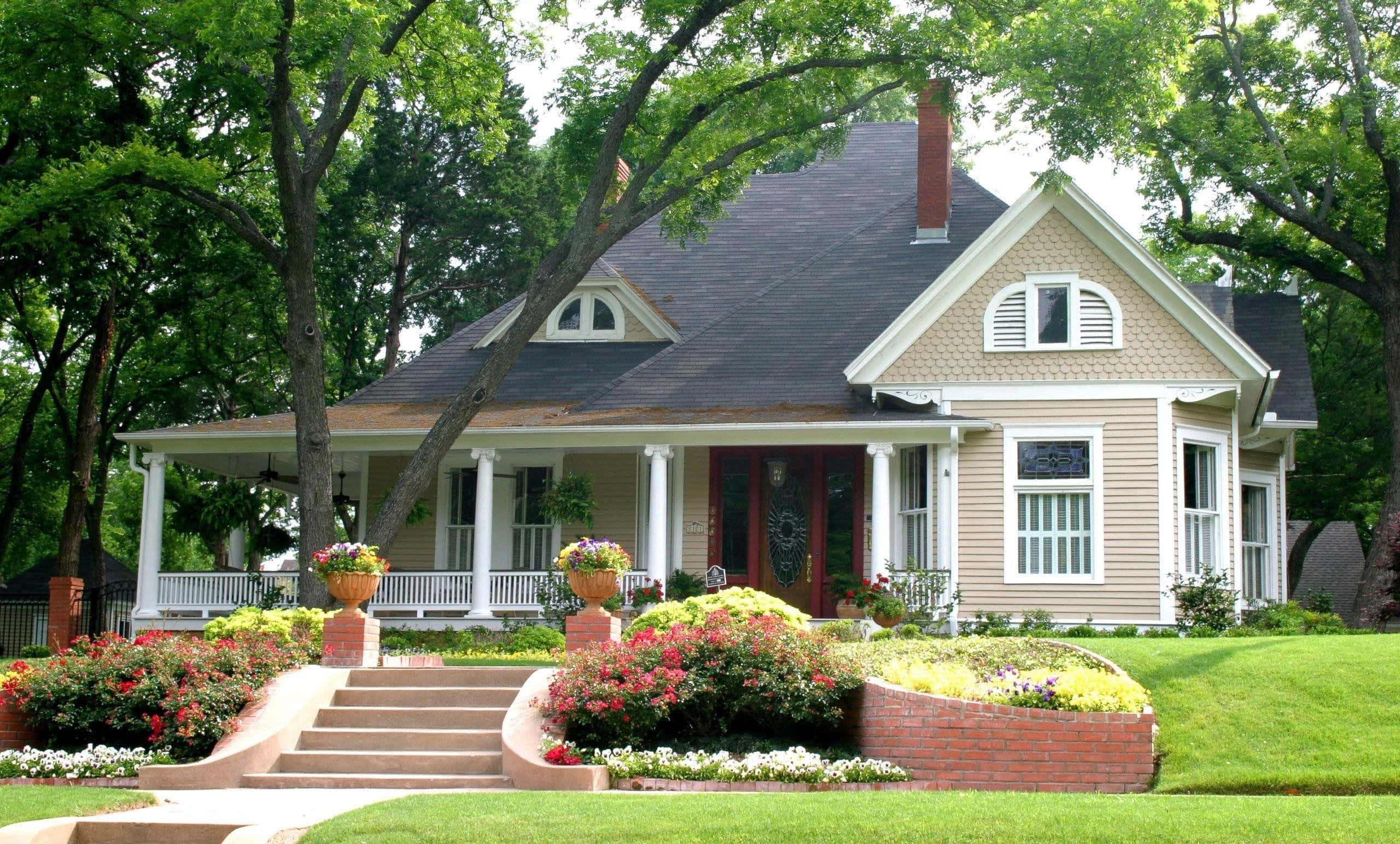 Beautiful House 桌布 壁紙 — 一座迷人的米色維多利亞風格房屋，帶有白色飾邊和溫馨的門廊，周圍環繞著茂密的綠色樹木和充滿活力的花壇。