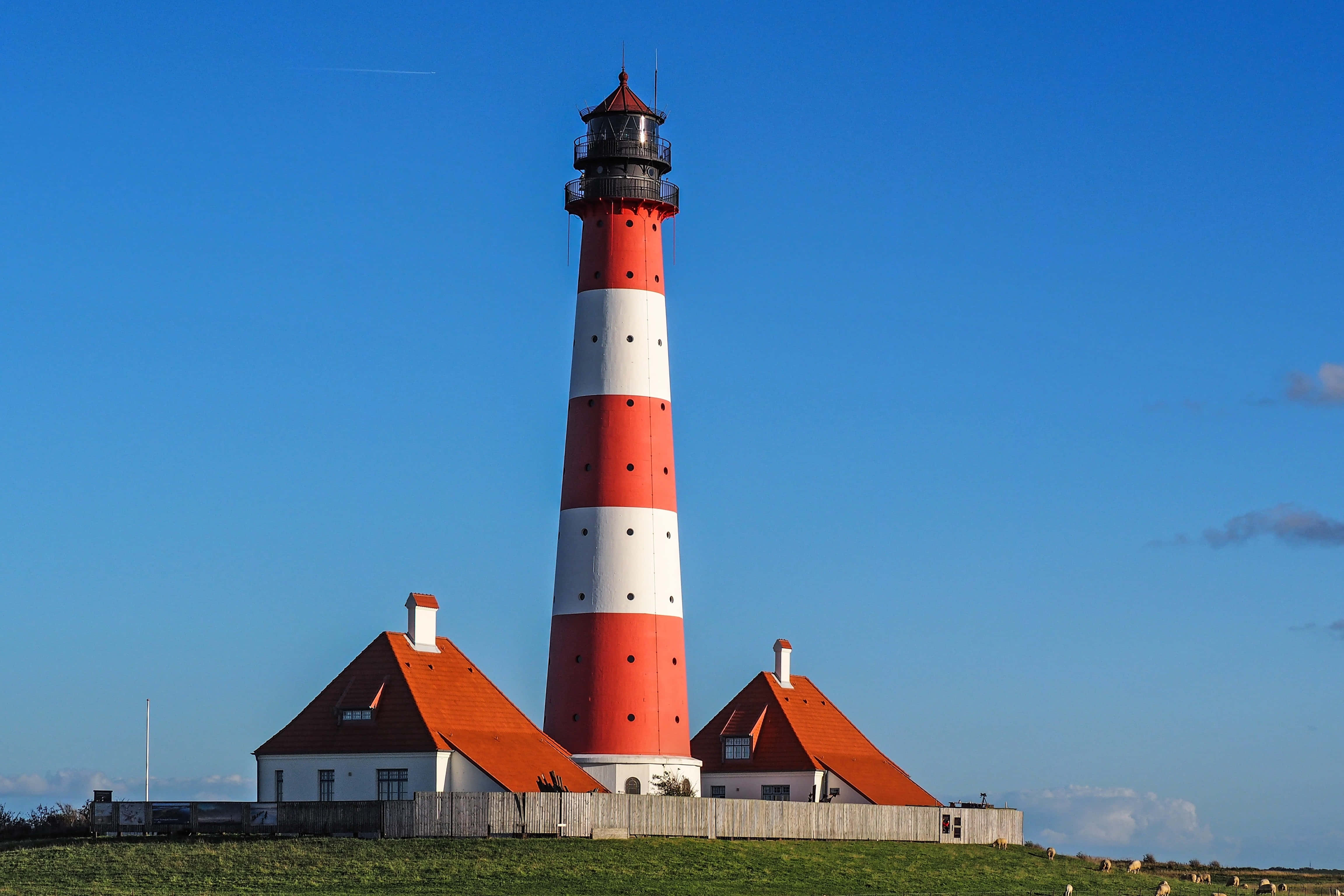 Lighthouse 桌布 壁紙 — 一座醒目的紅白條紋燈塔聳立在青草山坡上，藍天白雲下，附近有兩座小紅頂建築。