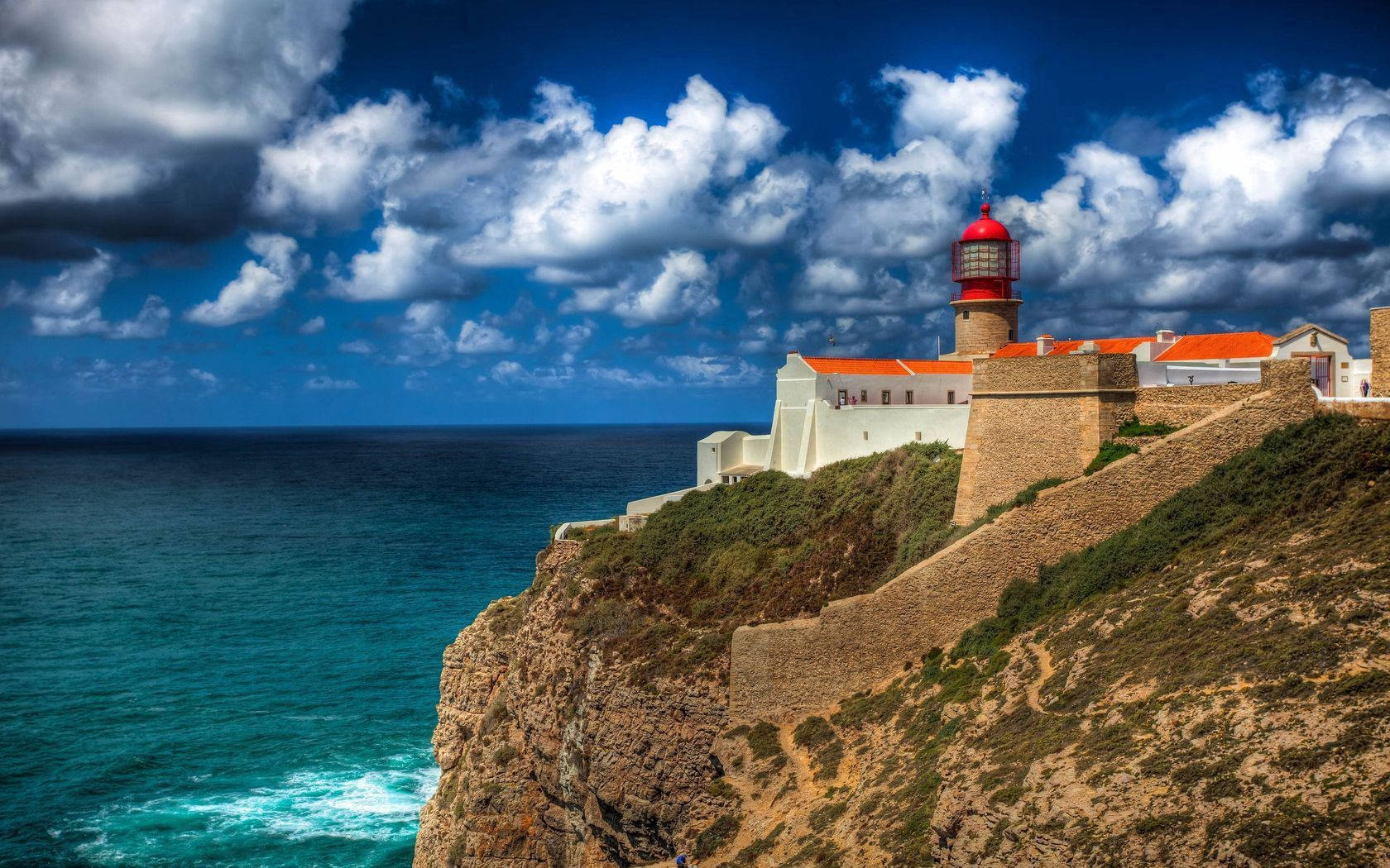 Portugal 桌布 壁紙 — 一座醒目的紅白燈塔矗立在崎嶇的懸崖頂上，俯瞰著碧綠的海浪，在戲劇性的藍天和蓬鬆的雲朵下。