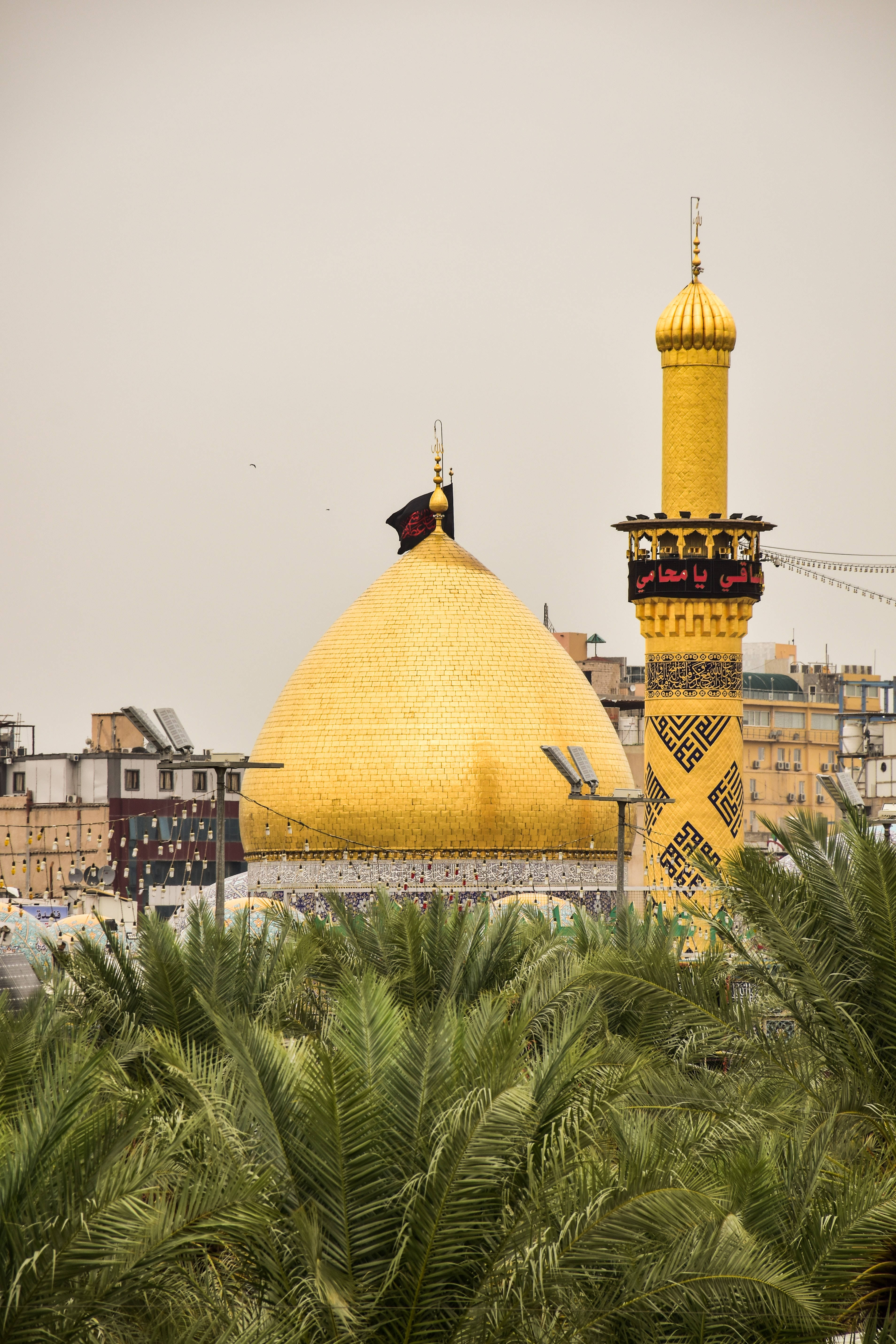Iraq 桌布 壁紙 — 一座金色圓頂清真寺，搭配相襯的宣禮塔，在朦朧的天空下聳立於茂密的棕櫚樹之上。
