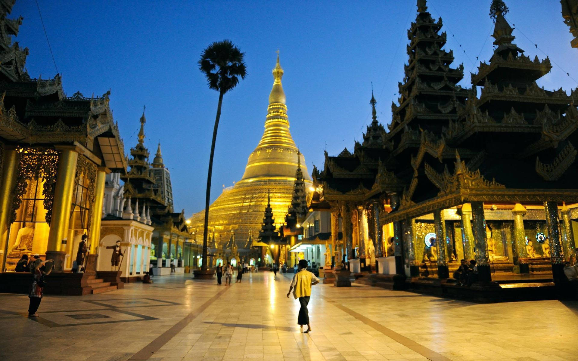 Yangon 桌布 壁紙 — 一座金色寶塔在暮色天空下溫暖地發光，一個孤獨的身影走過宏偉的寺廟庭院。