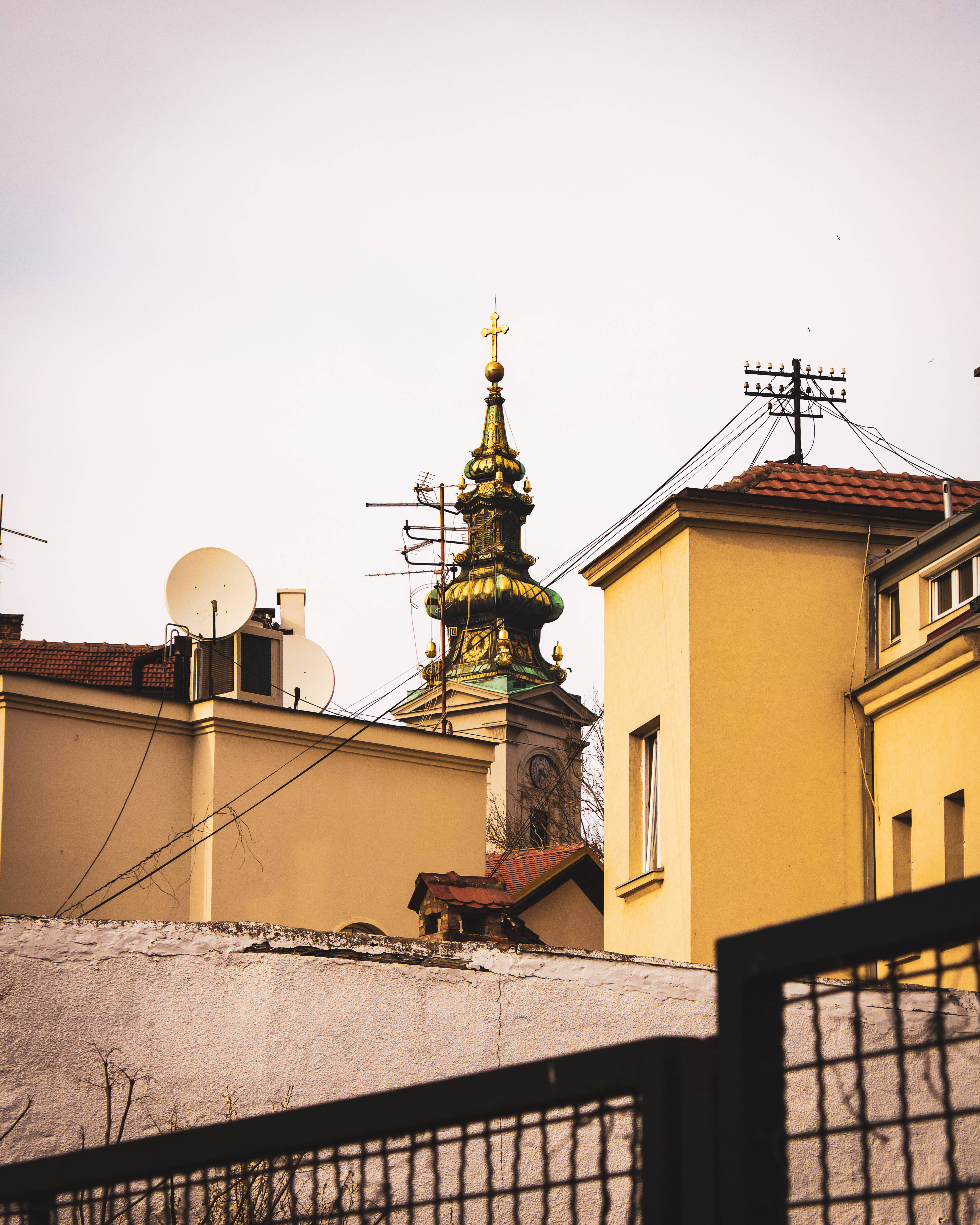 Serbia 桌布 壁紙 — 一座金色教堂尖塔在蒼白天空下，聳立於黃色建築物和纏繞的電線之上。
