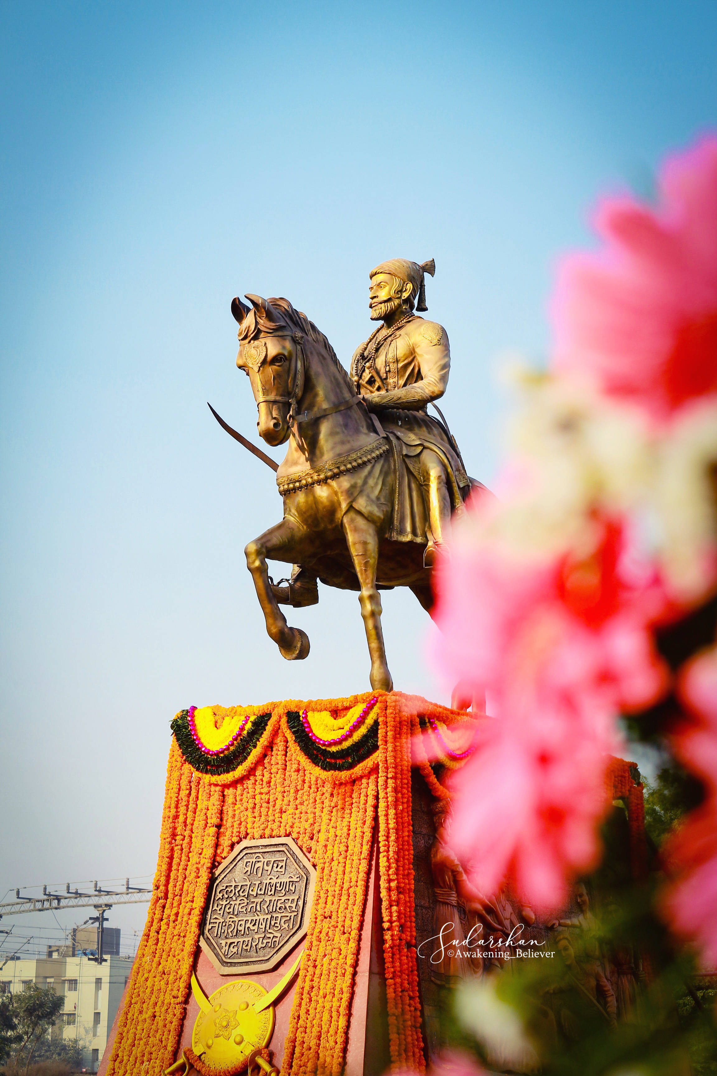 Indian 桌布 壁紙 — 一座金色的戰士騎馬雕像，馬匹後腿站立，立於色彩繽紛的花卉底座上，背景是清澈的藍天，前景有粉紅色的花朵。