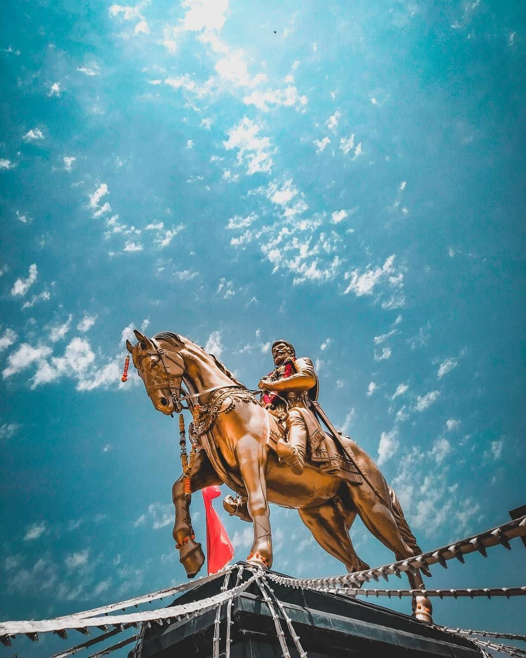 Indian 桌布 壁紙 — 一座金色的戰士騎馬雕像，騎著一匹後腿站立的馬，矗立在建築物頂部，背景是湛藍的天空和散落的白色雲朵。