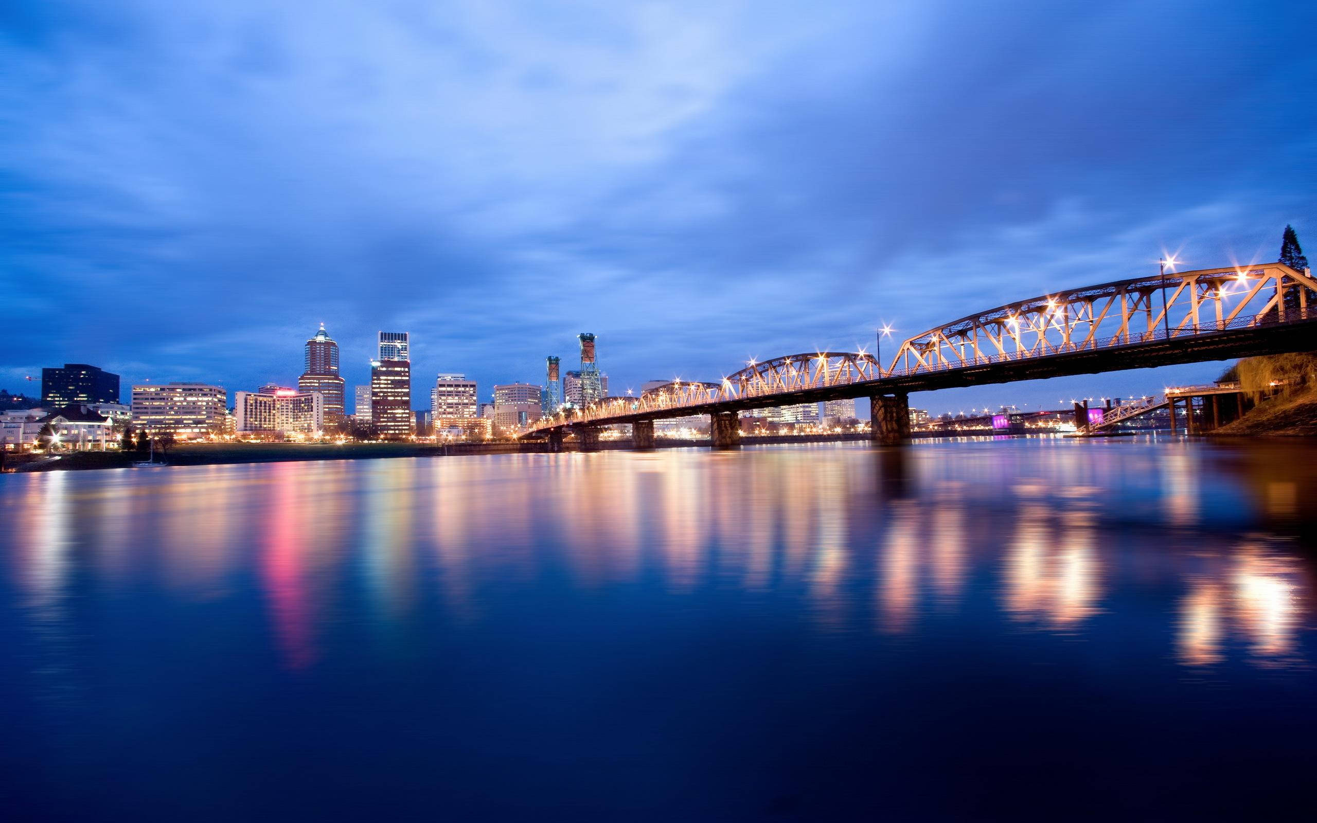 Portland 桌布 壁紙 — 一座鋼橋在暮色天空下拱跨一條平靜的河流，河面倒映著城市燈光。