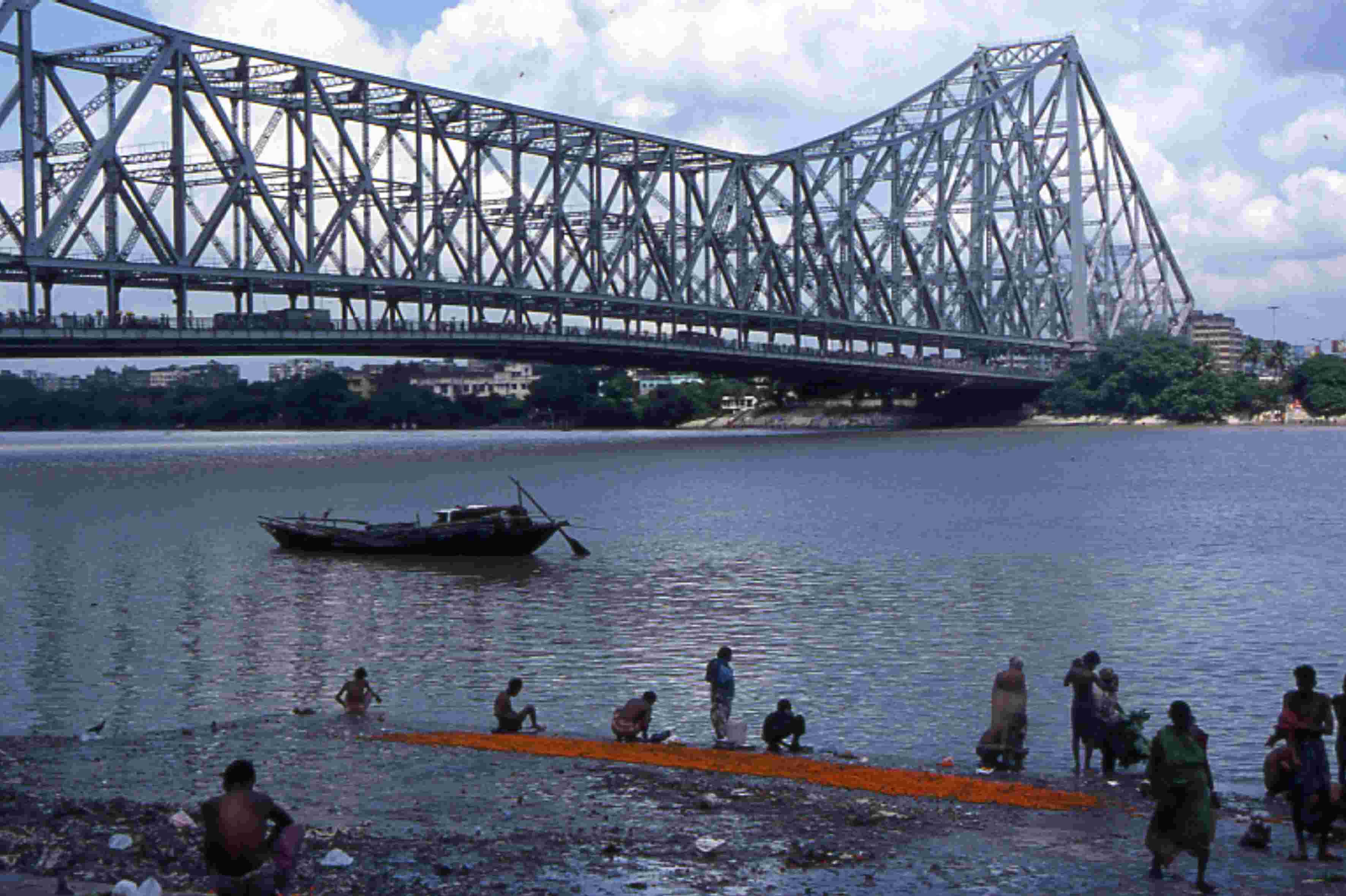 Kolkata 桌布 壁紙 — 一座鋼製桁架橋橫跨寬闊的河流，人們聚集在泥濘的河岸旁，旁邊有一艘小船。
