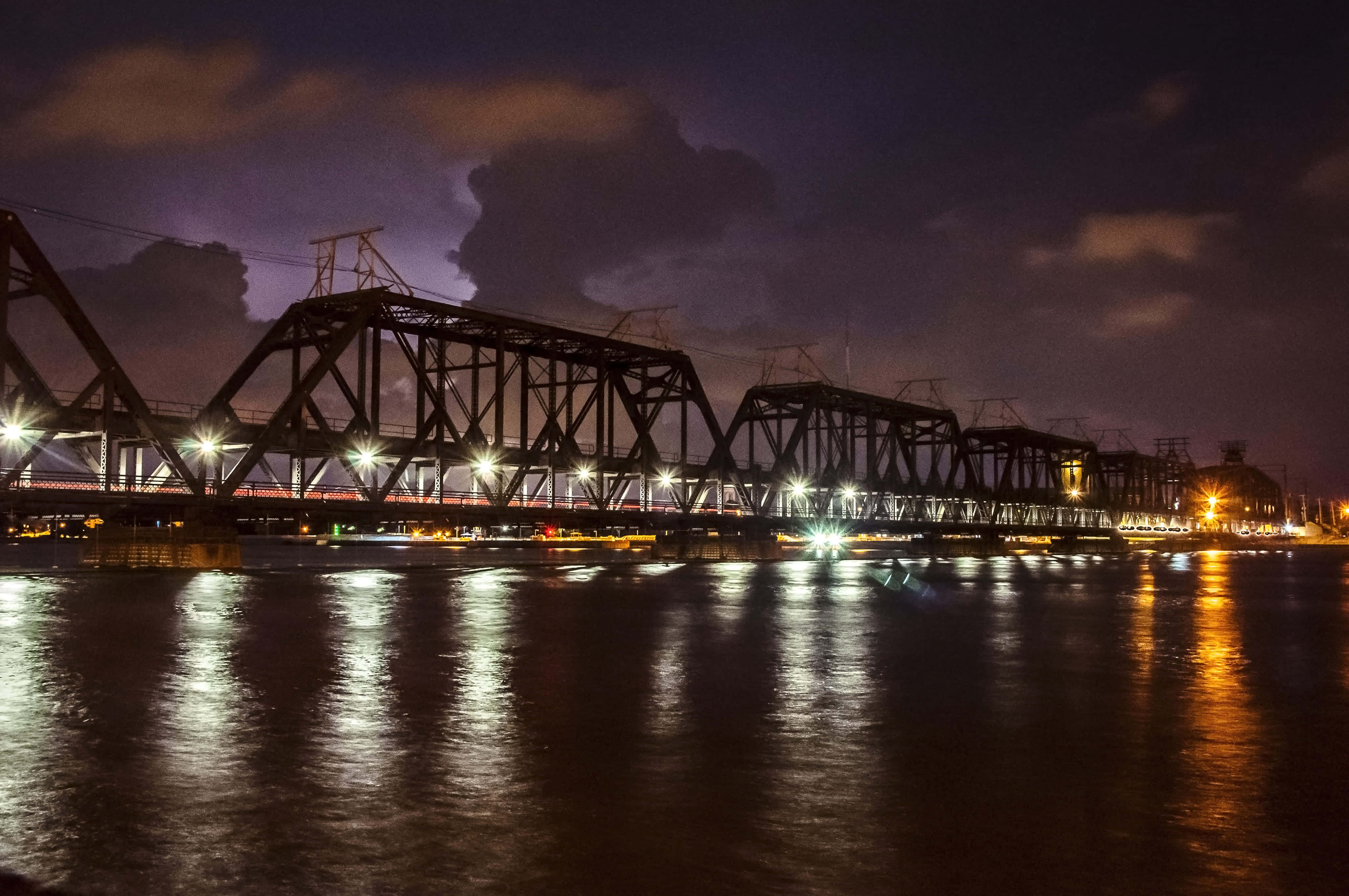 Louisiana 桌布 壁紙 — 一座鋼鐵桁架橋在陰鬱的紫色夜空下，在黑暗的河流上散發著溫暖的光芒。