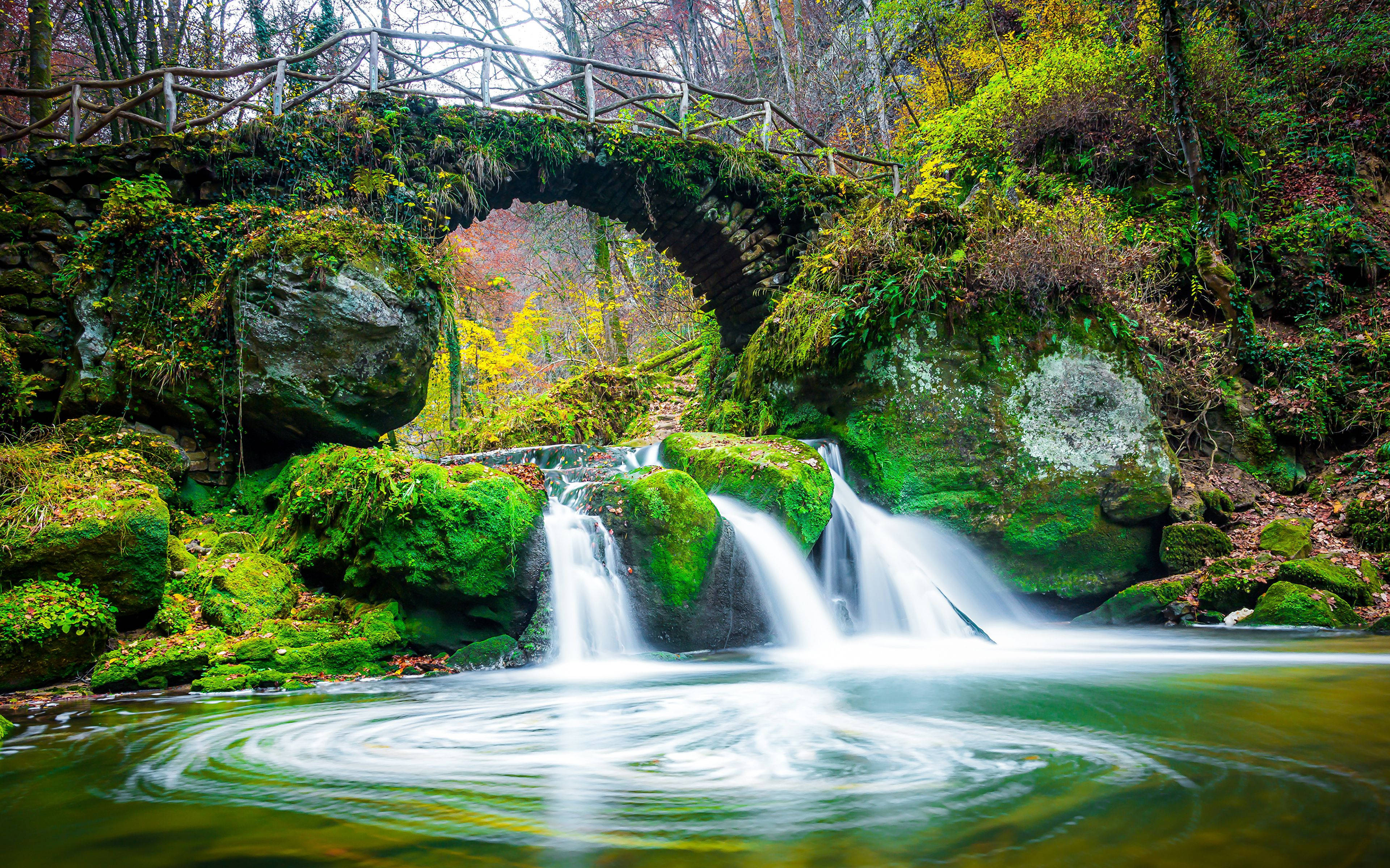 Luxembourg 桌布 壁紙 — 一座長滿青苔的石橋拱跨在瀑布之上，周圍是色彩繽紛的秋日森林。
