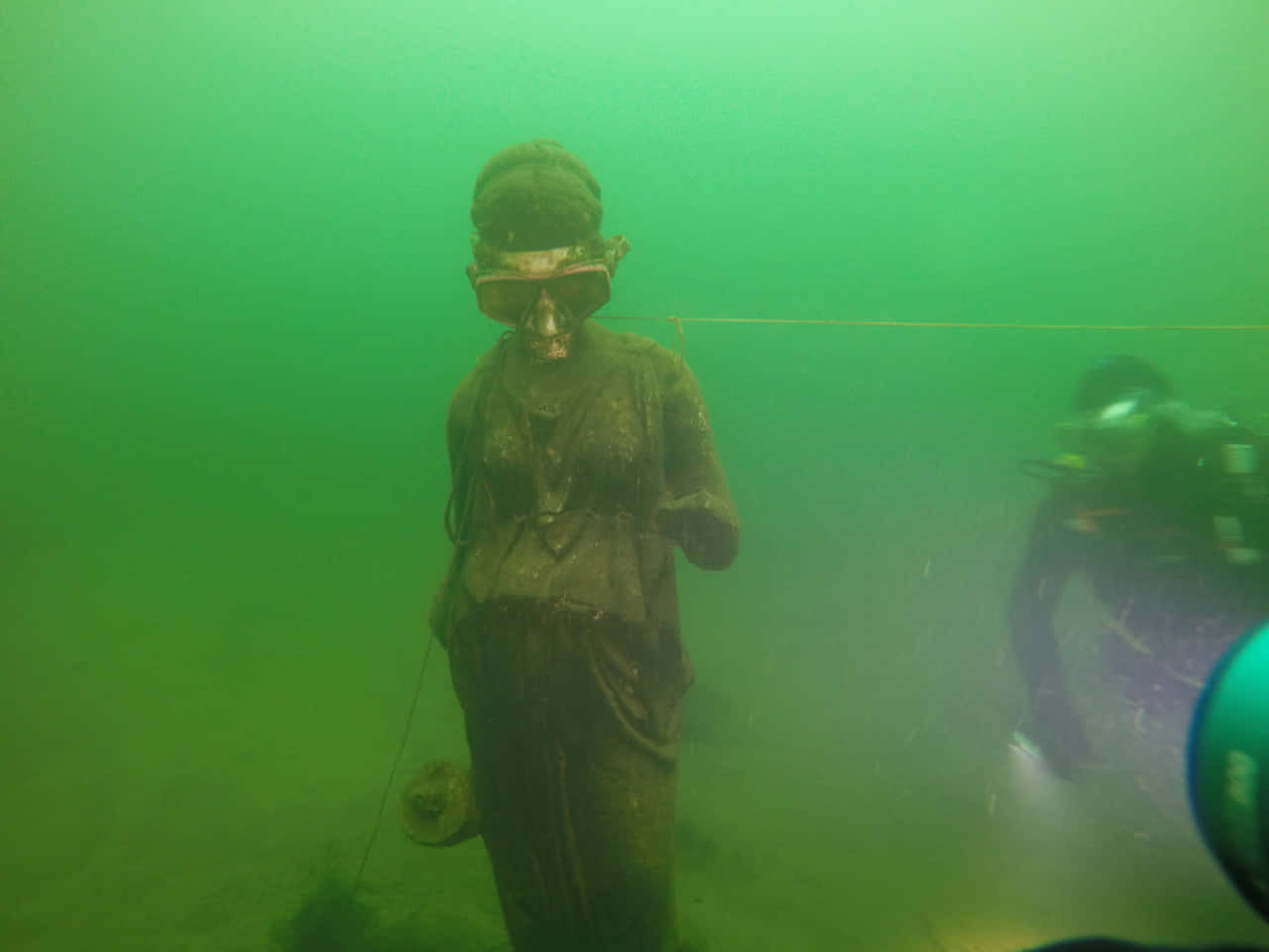 Scary Underwater 桌布 壁紙 — 一座長滿青苔的雕像浸在渾濁的綠色水中，附近有潛水員在探索。