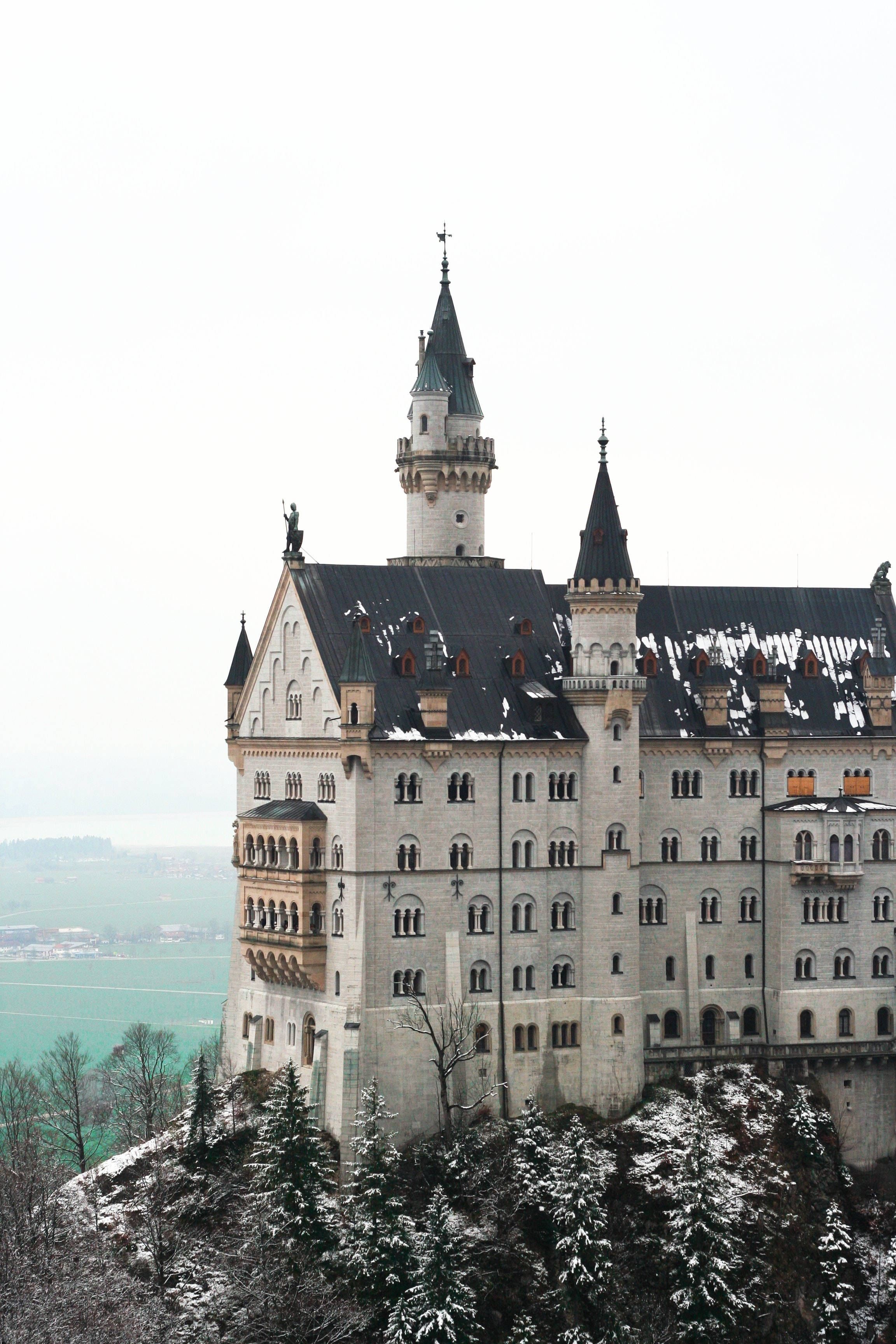 Neuschwanstein Castle 桌布 壁紙 — 一座雄偉的、覆蓋著白雪的童話城堡坐落在山頂上，下方是常青樹，遠處是朦朧的山谷。