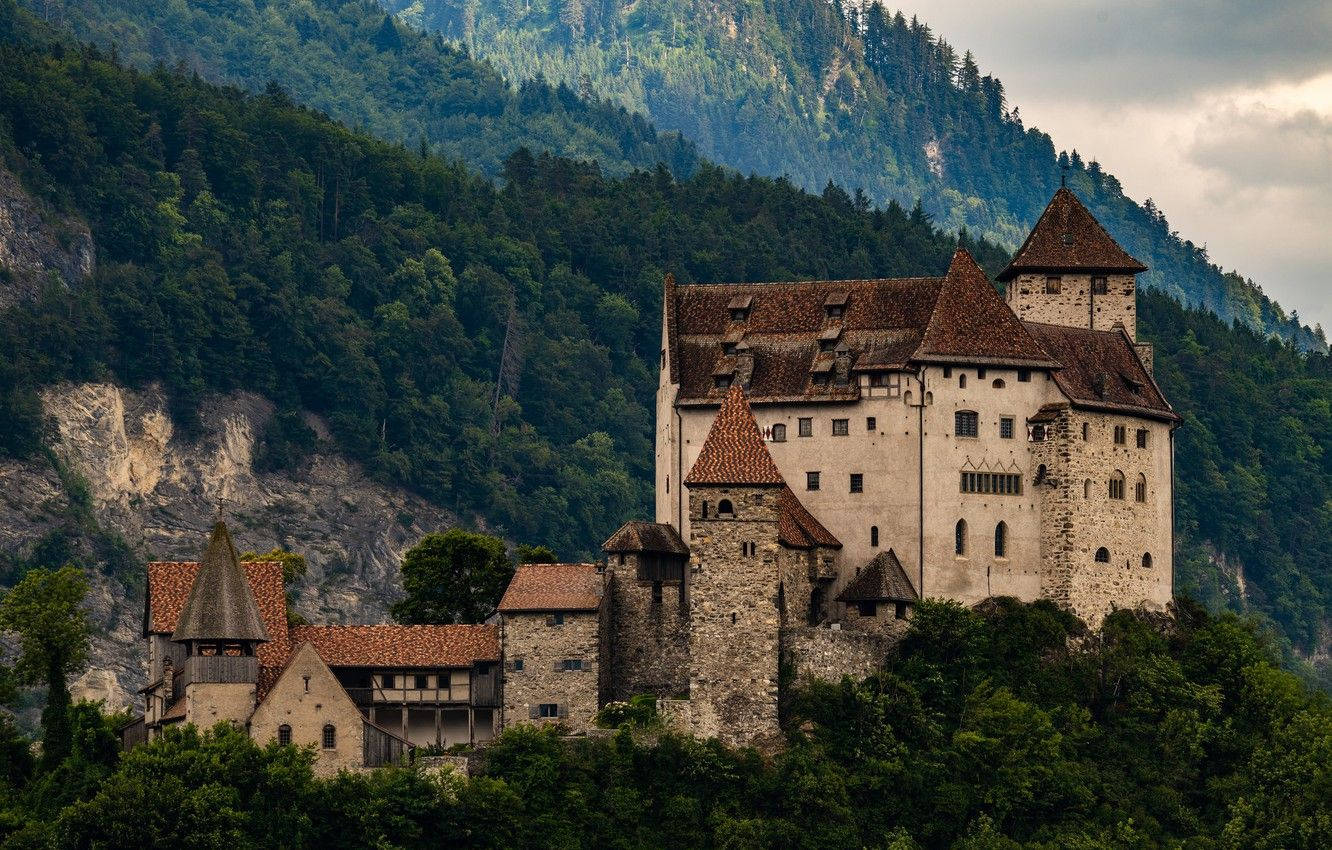 Liechtenstein 桌布 壁紙 — 一座雄偉的中世紀城堡，擁有石塔和紅色屋頂，緊貼在綠意盎然的山坡上，天空多雲。