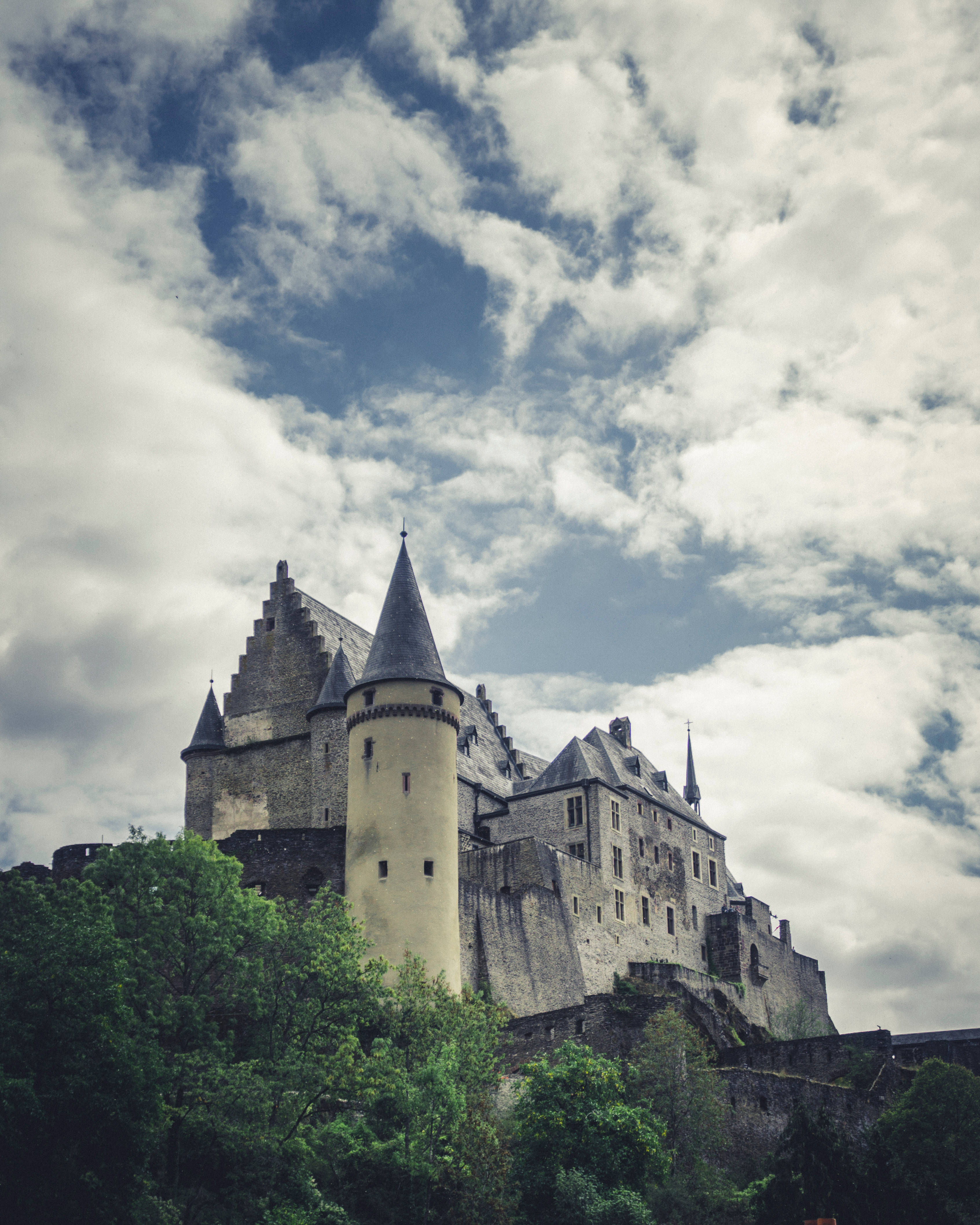 Luxembourg 桌布 壁紙 — 一座雄偉的中世紀城堡，擁有石塔和圓錐形屋頂，在戲劇性的多雲天空下，聳立在茂密的綠樹之上。