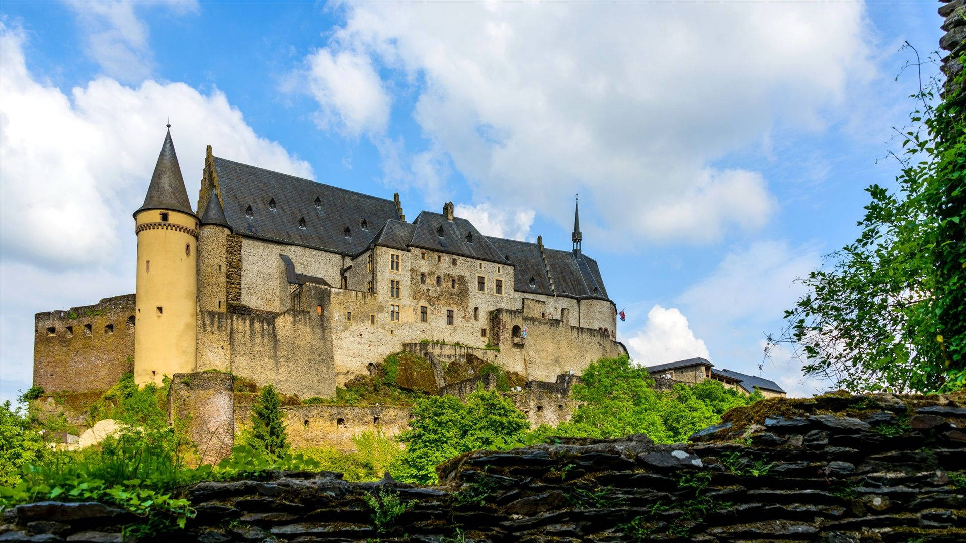 Luxembourg 桌布 壁紙 — 一座雄偉的中世紀城堡，擁有石塔和深色屋頂，坐落在山頂上，背景是明亮的藍天和蓬鬆的白雲。