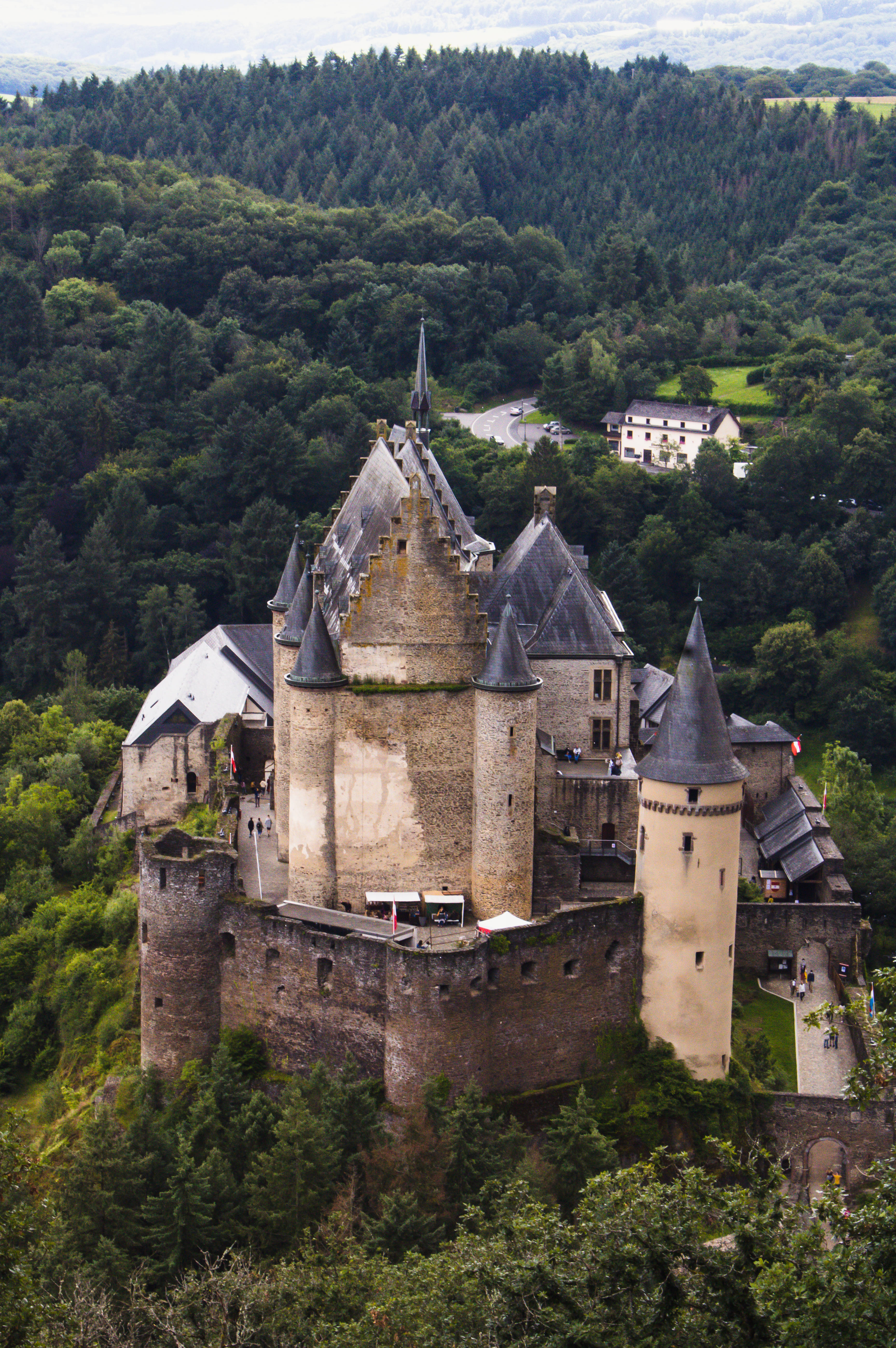 Luxembourg 桌布 壁紙 — 一座雄偉的中世紀城堡，擁有石塔和深色屋頂，坐落在森林覆蓋的山丘上，俯瞰著散落著房屋的山谷。