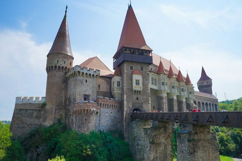 Romania 桌布 壁紙 — 一座雄偉的中世紀城堡，擁有紅色瓦片屋頂和石塔，矗立在鬱鬱蔥蔥的綠色山丘上，天空湛藍明亮。