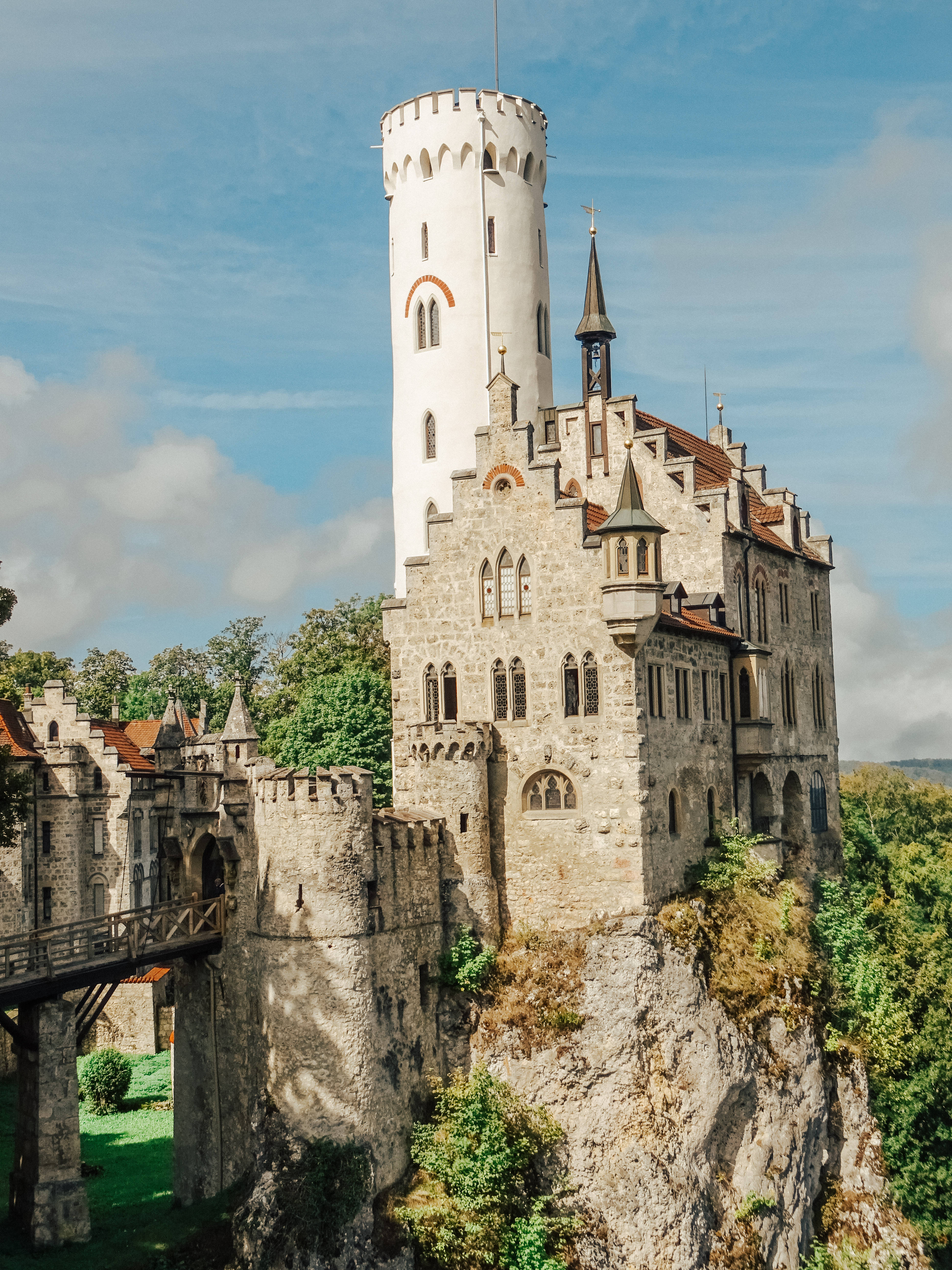 Liechtenstein 桌布 壁紙 — 一座雄偉的中世紀城堡坐落在岩石懸崖上，周圍綠樹成蔭，有一座木橋，在明亮的藍天下。