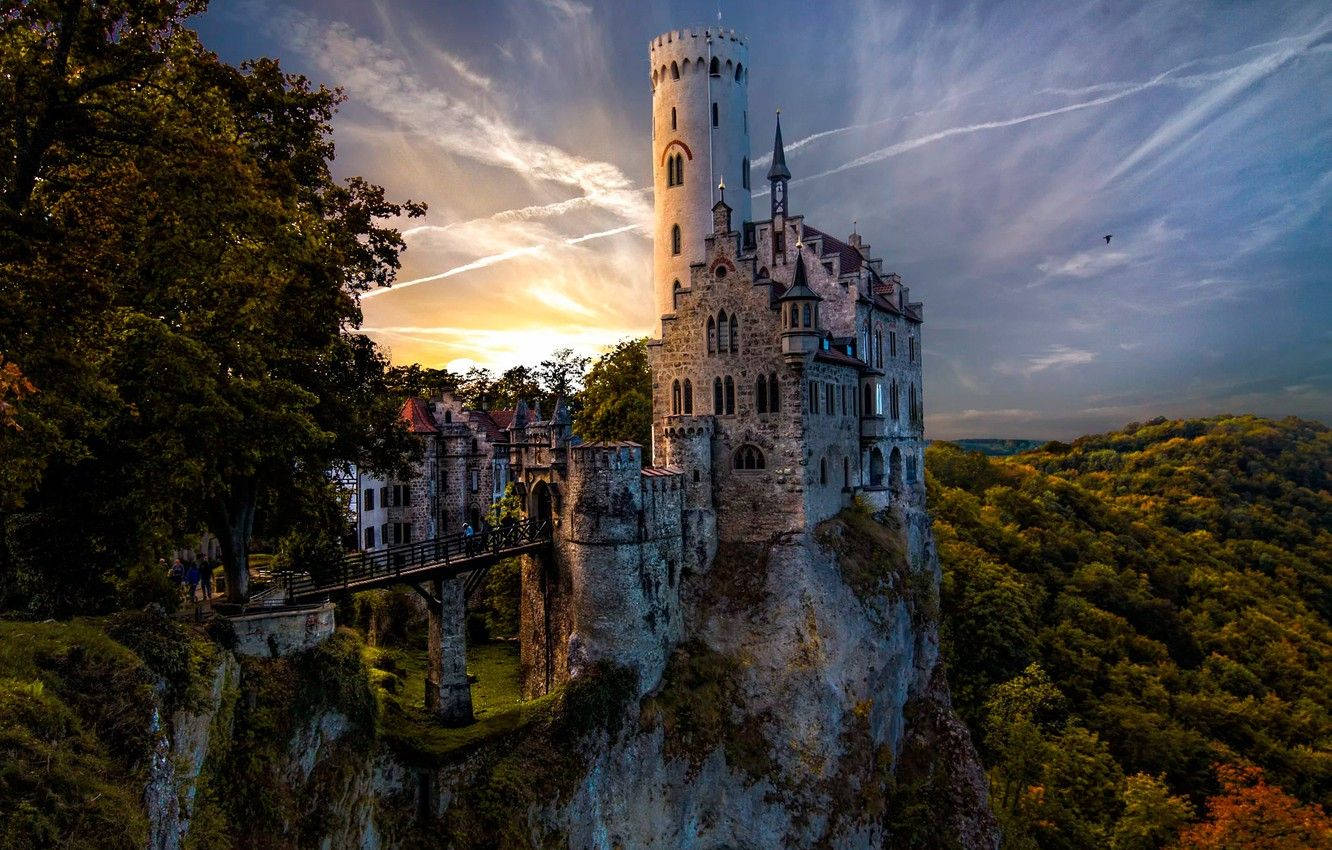 Liechtenstein 桌布 壁紙 — 一座雄偉的中世紀城堡坐落在懸崖上，夕陽西下，金色的光芒照亮了它的石塔和周圍的秋林。
