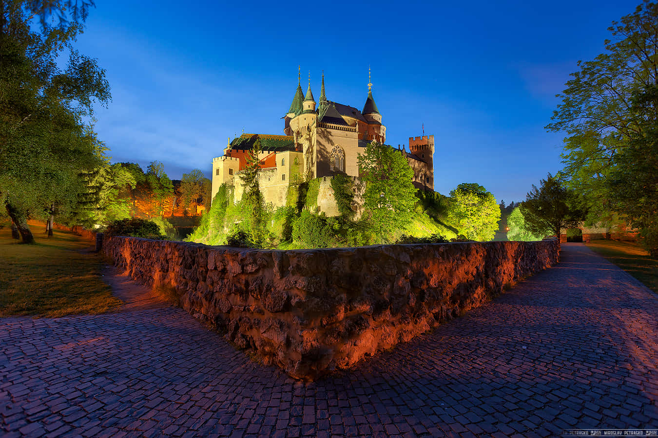 城堡動畫 桌布 壁紙 — 一座雄偉的中世紀城堡坐落在暮色中的山丘上，溫暖的燈光照亮其石塔和周圍的綠植。