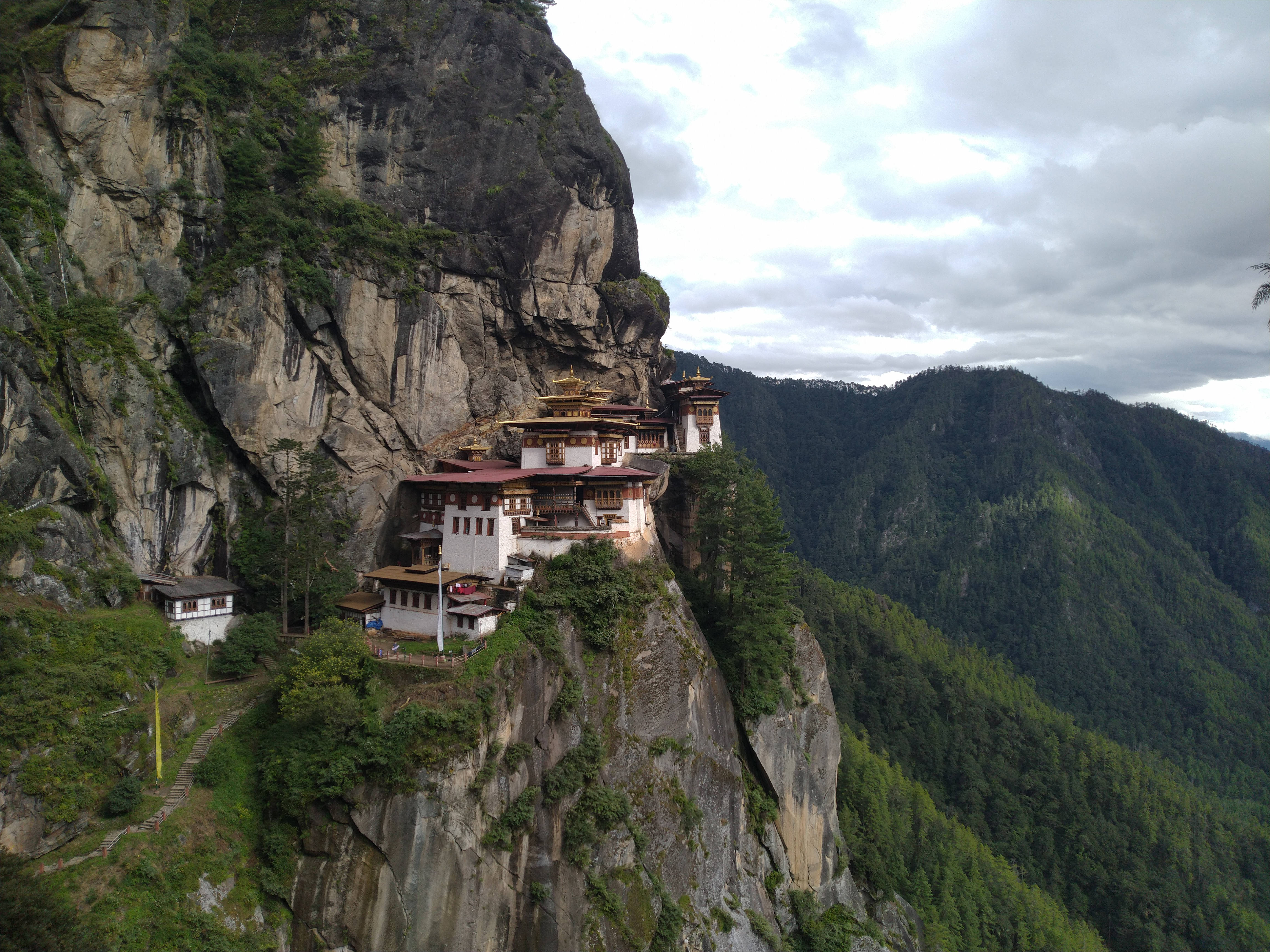 Bhutan 桌布 壁紙 — 一座雄偉的佛教寺院依附在陡峭的懸崖面上，周圍是鬱鬱蔥蔥的青山，天空多雲。