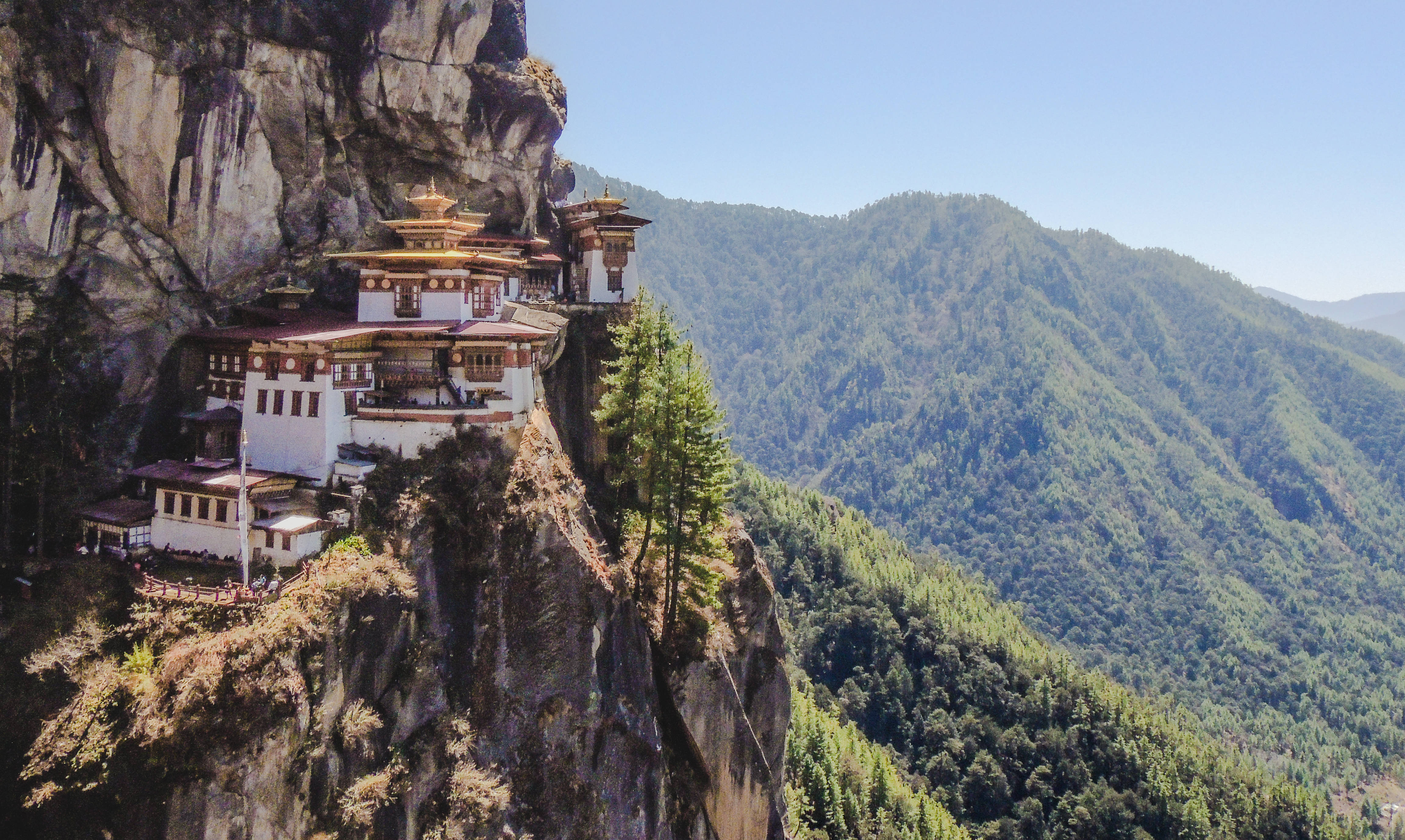 Bhutan 桌布 壁紙 — 一座雄偉的佛教寺院緊貼在陡峭的懸崖上，俯瞰著蔚藍天空下鬱鬱蔥蔥的綠色山谷。