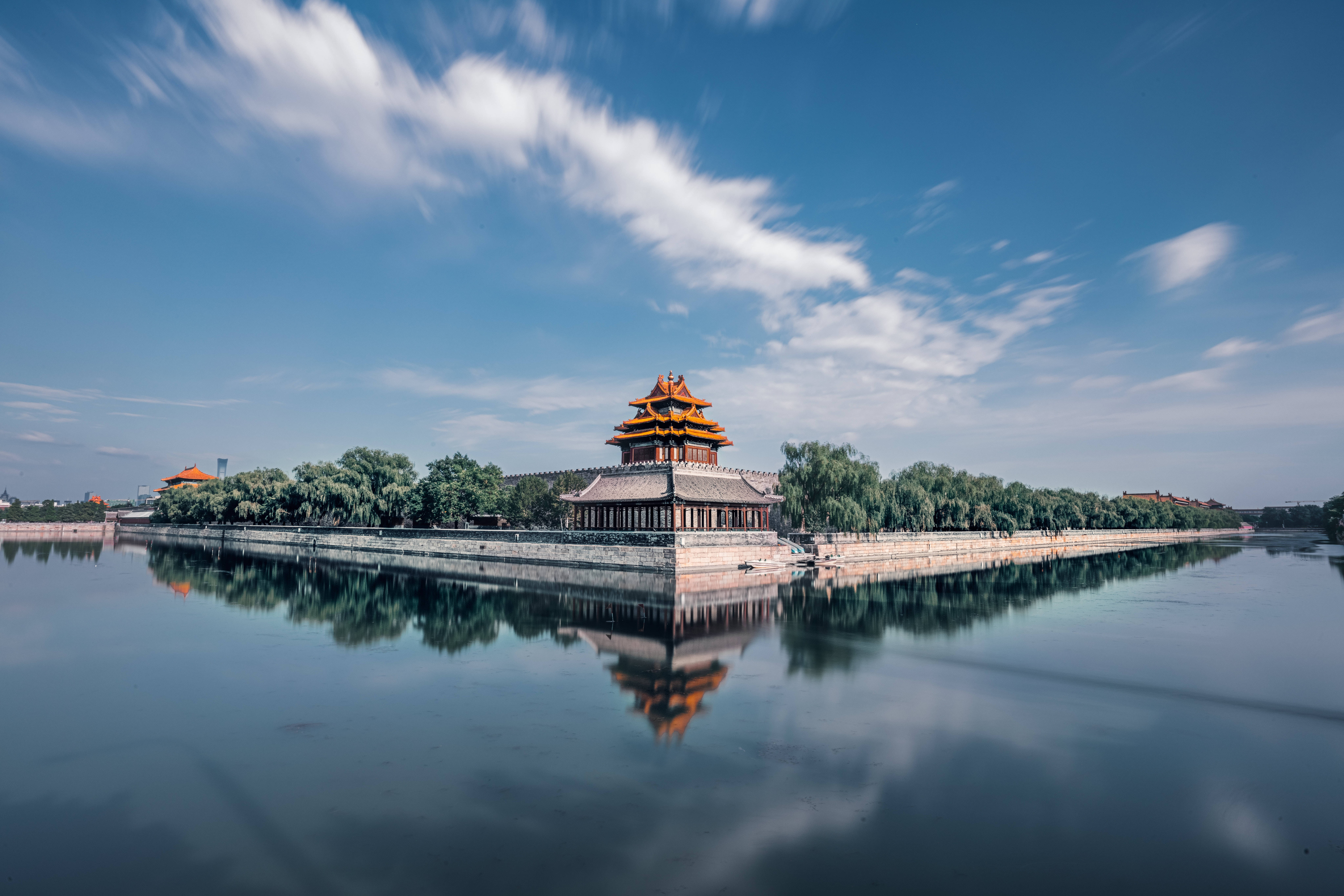 高清 桌布 壁紙 — 一座雄偉的傳統中式宮殿，擁有橙色屋頂和灰色牆壁，靜靜地矗立在石台上，在藍天白雲的映襯下，完美地倒映在平靜的水面上。
