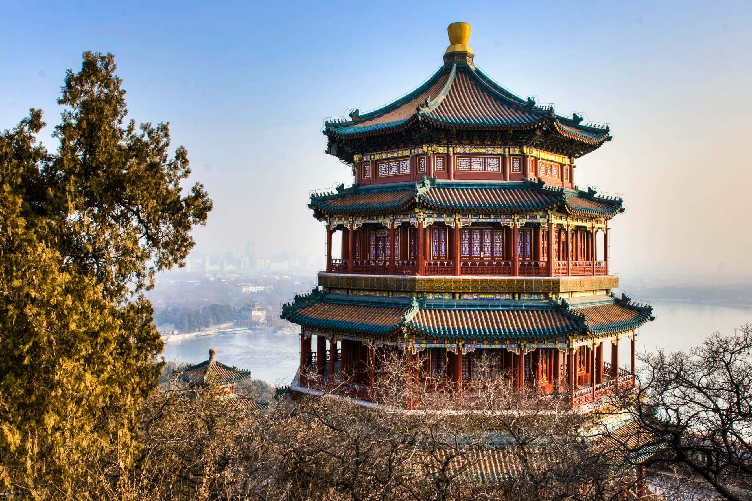 建築 桌布 壁紙 — 一座雄偉的傳統中式寶塔，紅色柱子和綠色瓦片屋頂，矗立在山丘上，俯瞰著寧靜的湖泊和遠處的城市景觀，天空清澈湛藍。