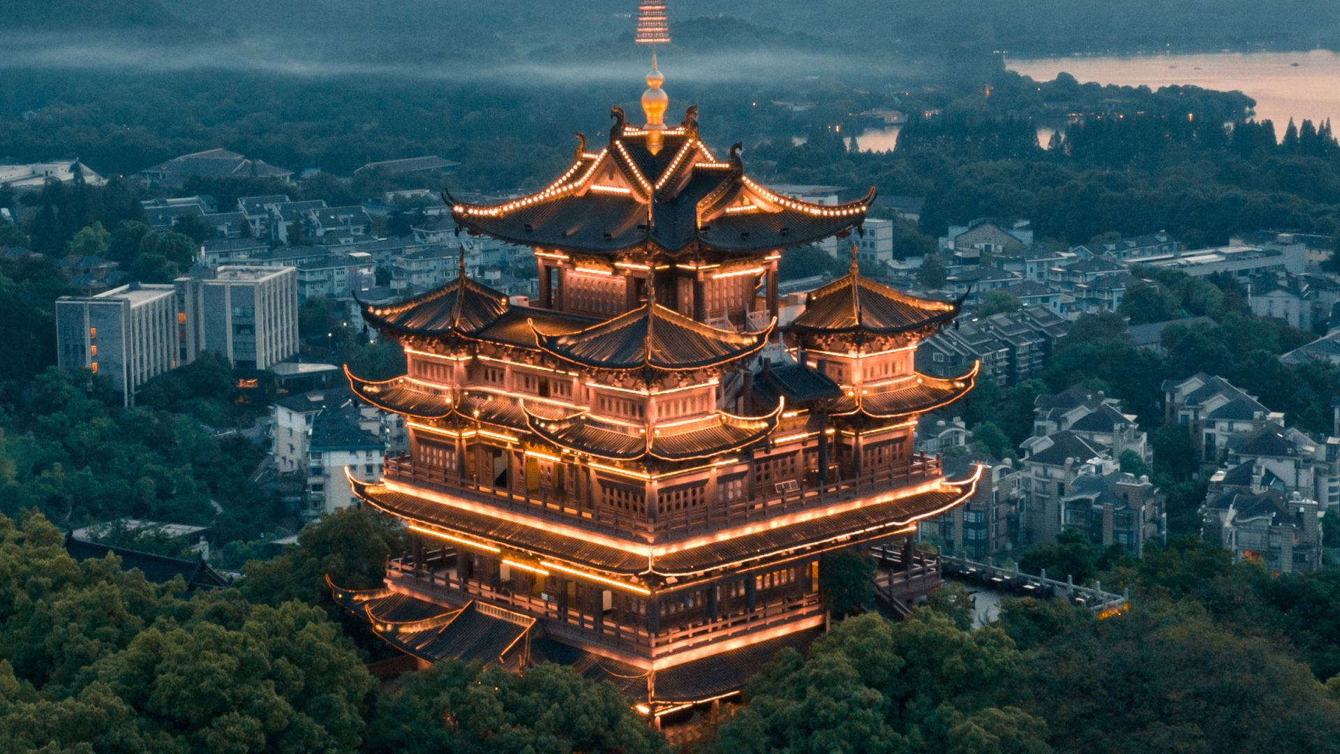 Hangzhou 桌布 壁紙 — 一座雄偉的傳統中式寶塔在燈光下溫暖地發光，映襯著霧氣瀰漫的暮色天空，下方是密集的城市景觀。