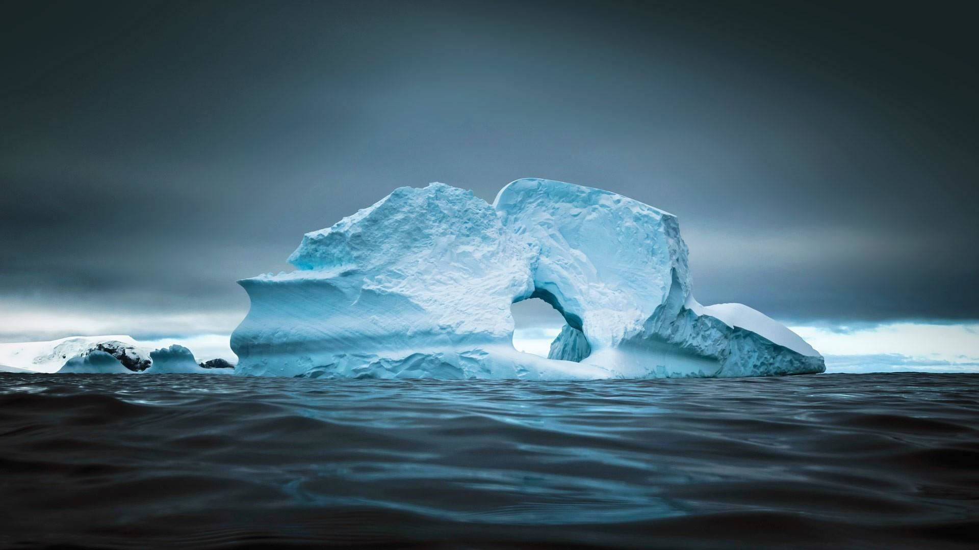 Antarctica 桌布 壁紙 — 一座雄偉的冰山帶有天然拱門，在陰沉灰暗的天空下靜靜漂浮在黑暗的水面上。