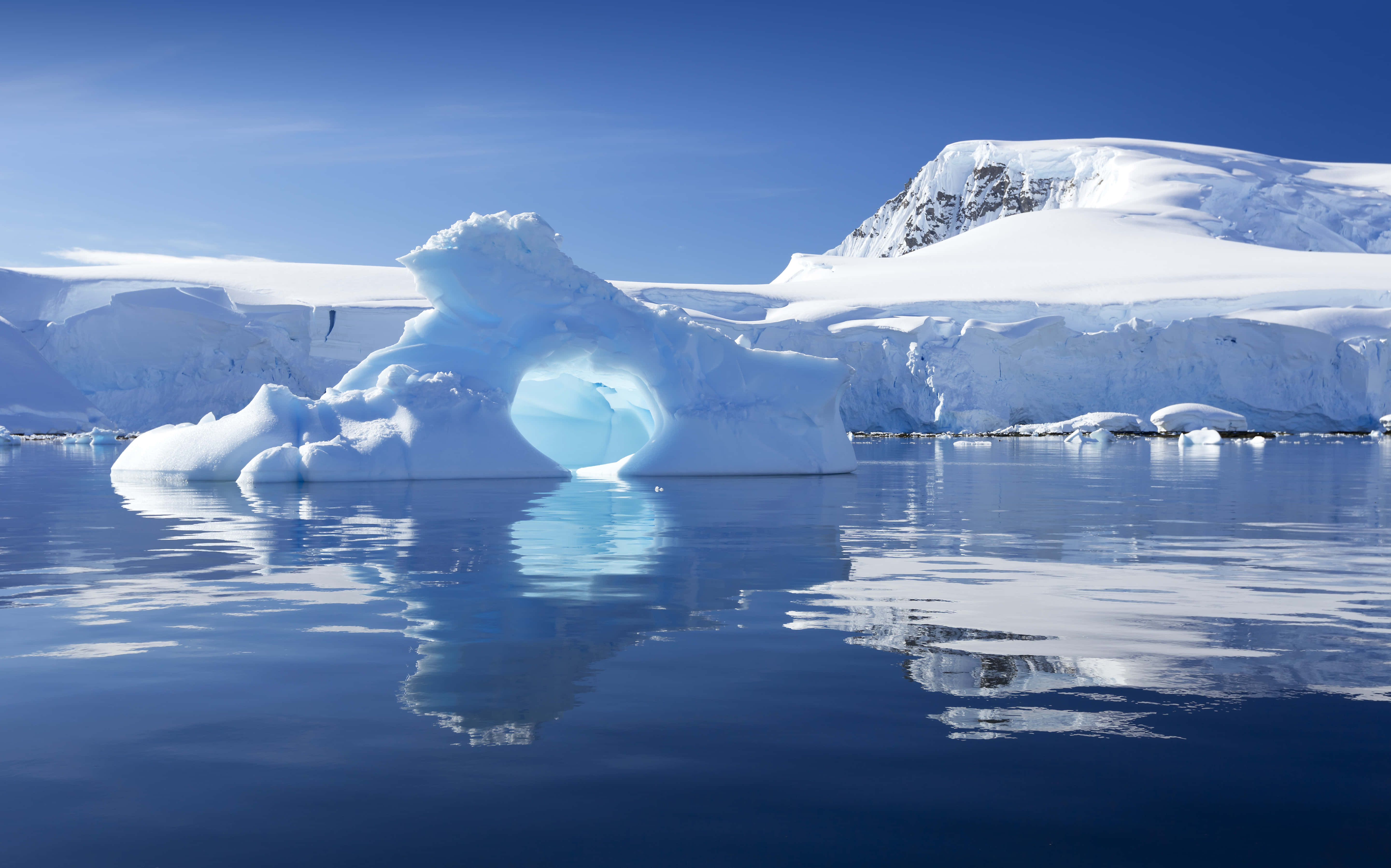 Antarctica 桌布 壁紙 — 一座雄偉的冰山帶有完美的拱門，在清澈的天空下寧靜地漂浮在深藍色的海水中，背景是白雪覆蓋的山脈。