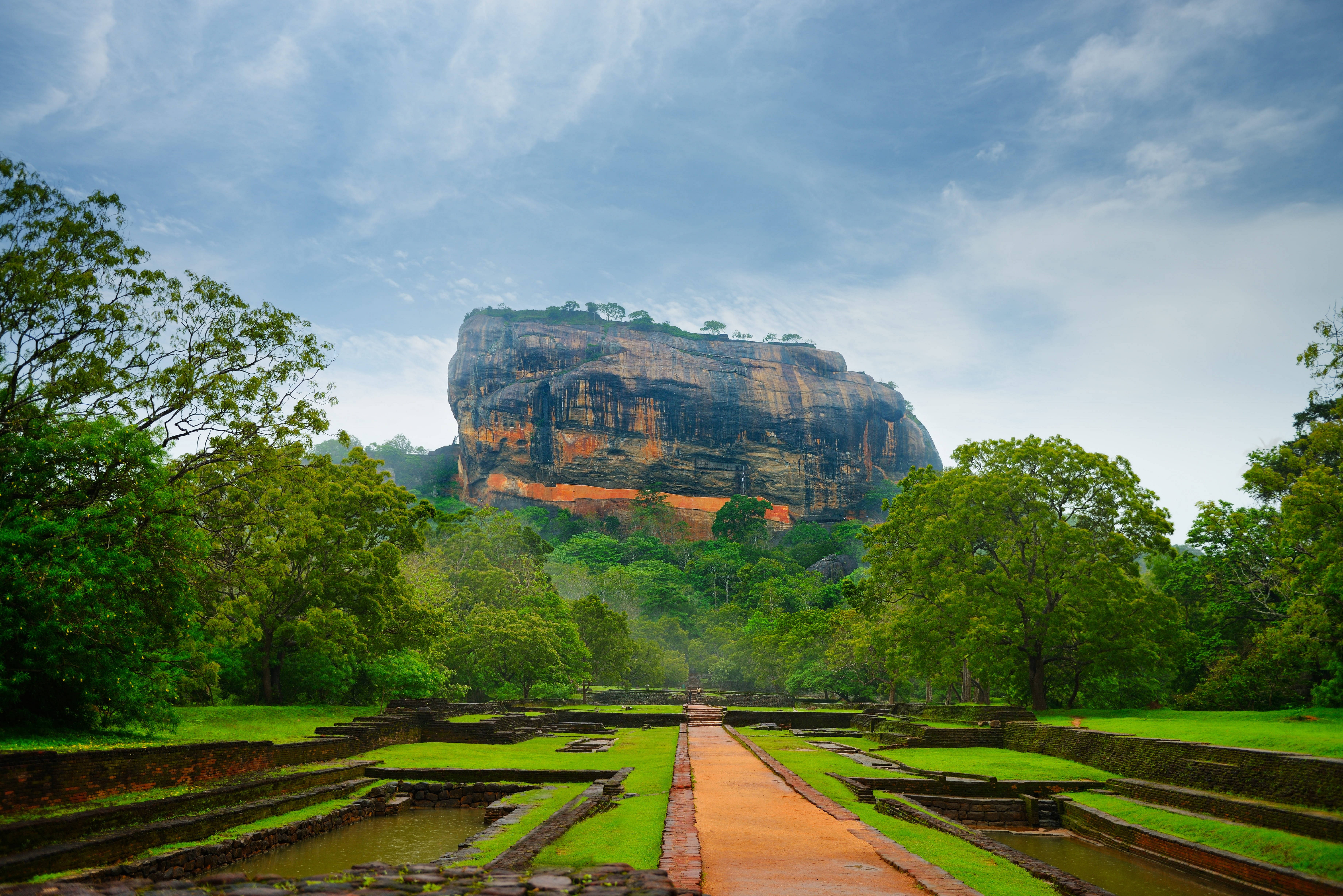 Sri Lanka 桌布 壁紙 — 一座雄偉的古老岩石堡壘在部分多雲的天空下，聳立在鬱鬱蔥蔥的綠色花園和石徑之上。