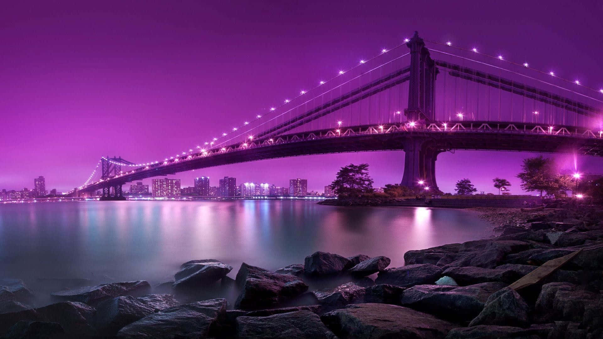 1920X1080 桌布 壁紙 — 一座雄偉的吊橋在夜晚發出紫色光芒，橫跨平靜的水面，前景是岩石。