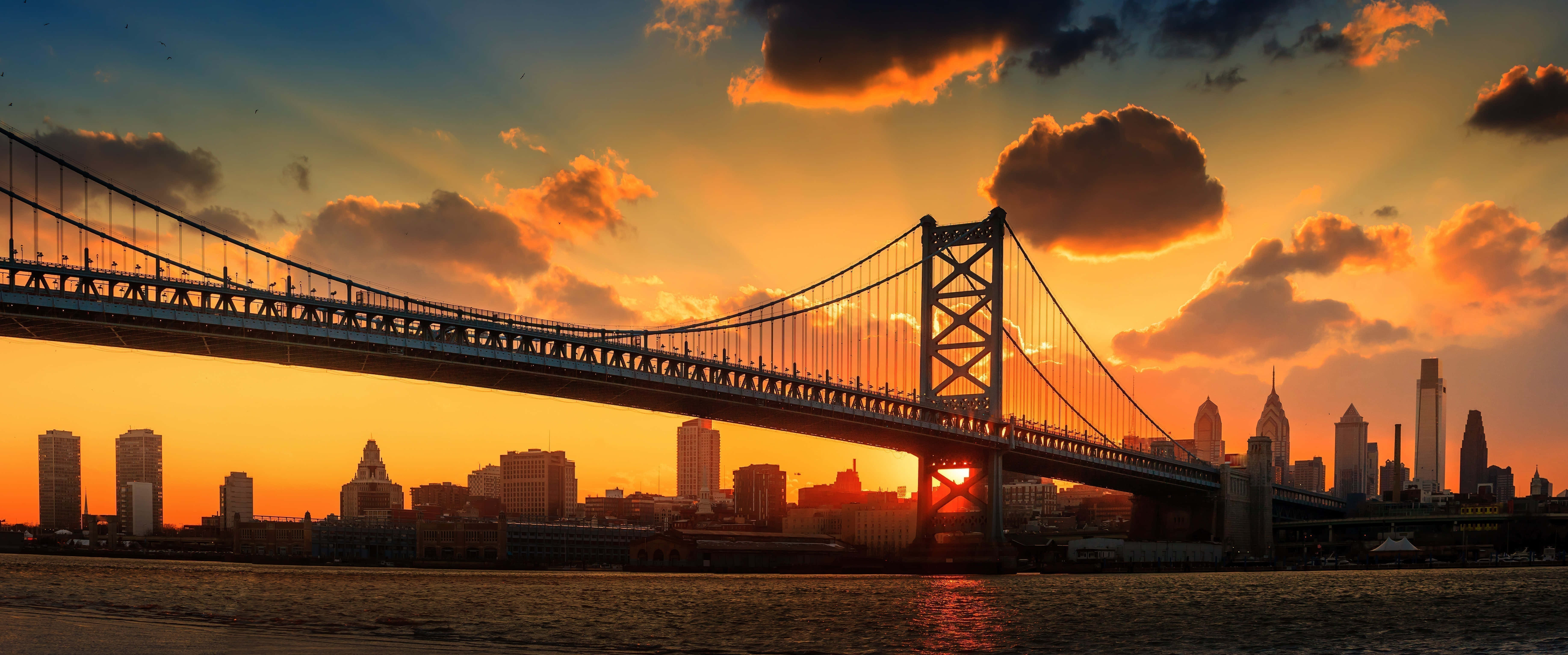 bridge 桌布 壁紙 — 一座雄偉的吊橋在日落時分橫跨河流，天空有火紅橙色的雲彩和城市天際線剪影。