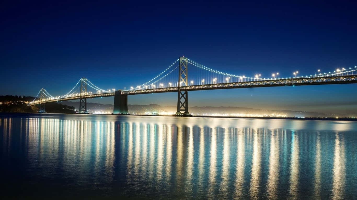 1366x768 San Francisco 桌布 壁紙 — 一座雄偉的吊橋在深藍色的夜空下散發著溫暖的光芒，其倒影在下方平靜的水面上閃爍。