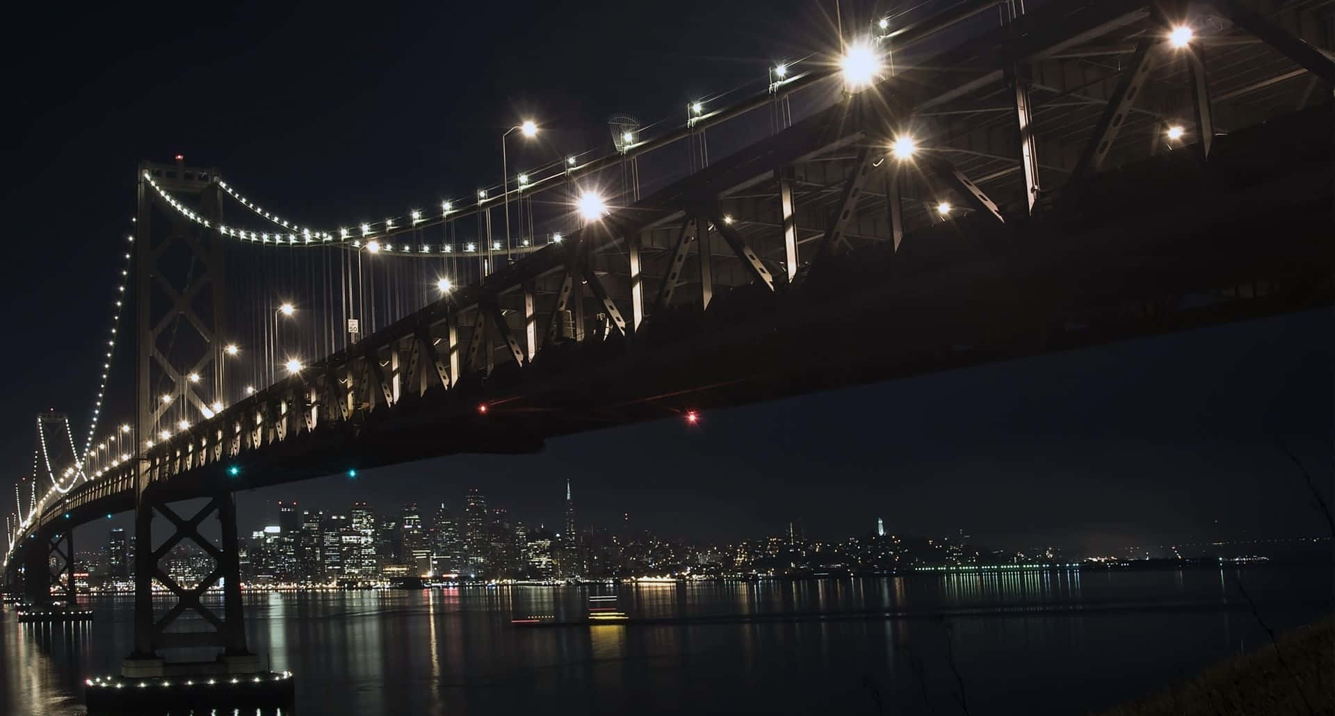 1920X1080 桌布 壁紙 — 一座雄偉的吊橋在漆黑的夜空中散發出溫暖的光芒，其倒影在下方平靜的水面上閃爍，遠處是充滿活力的城市天際線。