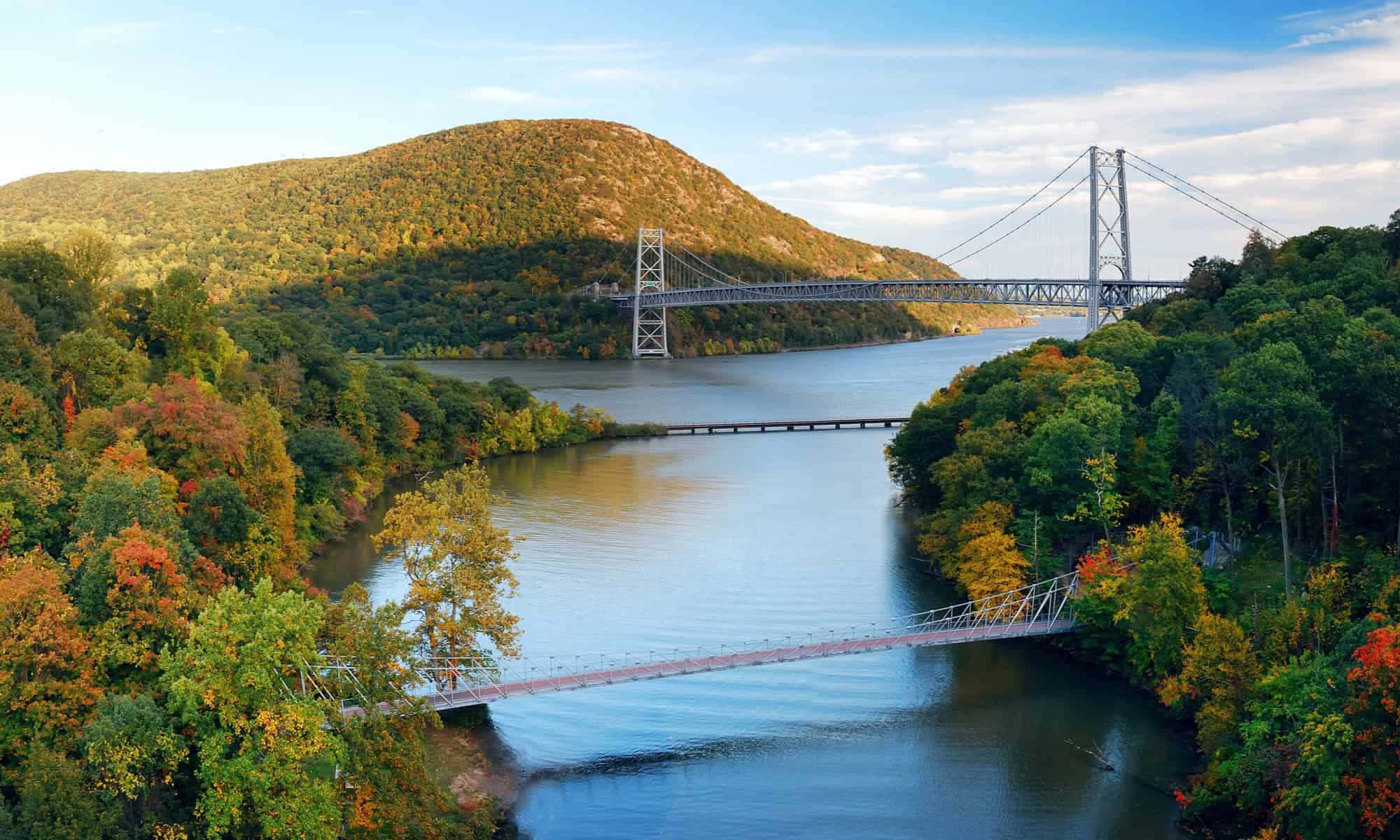 river 桌布 壁紙 — 一座雄偉的吊橋橫跨寬闊的河流，兩旁是秋日森林和起伏的山丘，上方是明亮的藍天。