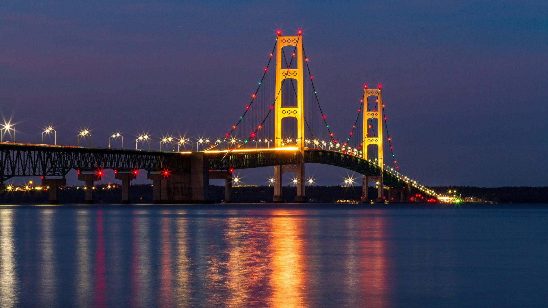 夜空 桌布 壁紙 — 一座雄偉的吊橋裝飾著節慶燈光，在暮色中平靜的水面上閃耀。