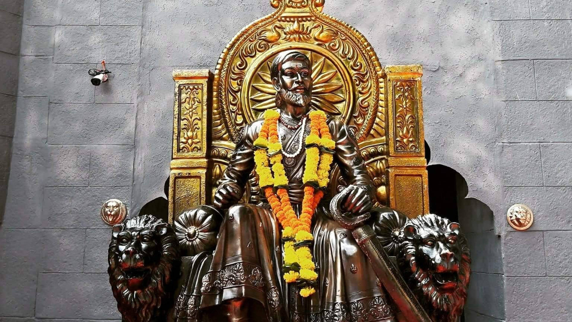 希瓦吉 Maharaj 高清壁紙 桌布 壁紙 — 一座雄偉的國王青銅雕像，鬍鬚濃密，坐在金色寶座上，兩側有獅頭裝飾，並飾以鮮豔的黃色和橙色萬壽菊花環。