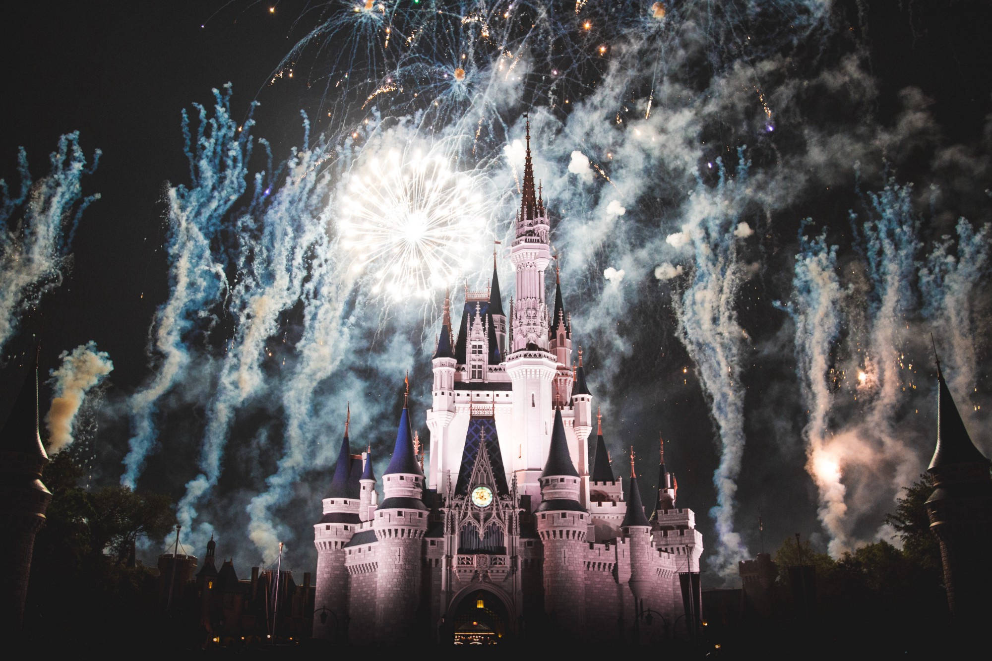 Disneyland Desktop 桌布 壁紙 — 一座雄偉的城堡在夜晚被照亮，鮮豔的藍色和白色煙火在其塔樓上方爆炸。