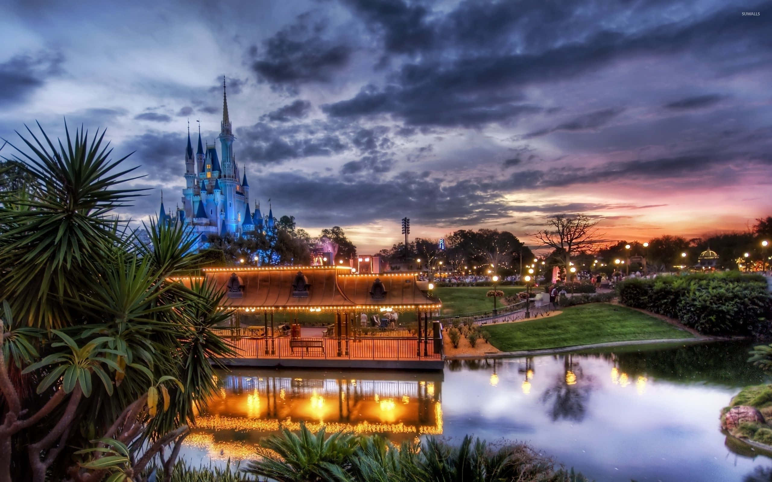 Disney World Desktop 桌布 壁紙 — 一座雄偉的城堡在暮色天空下閃耀，其燈光倒映在寧靜的池塘中，四周環繞著茂密的綠植。