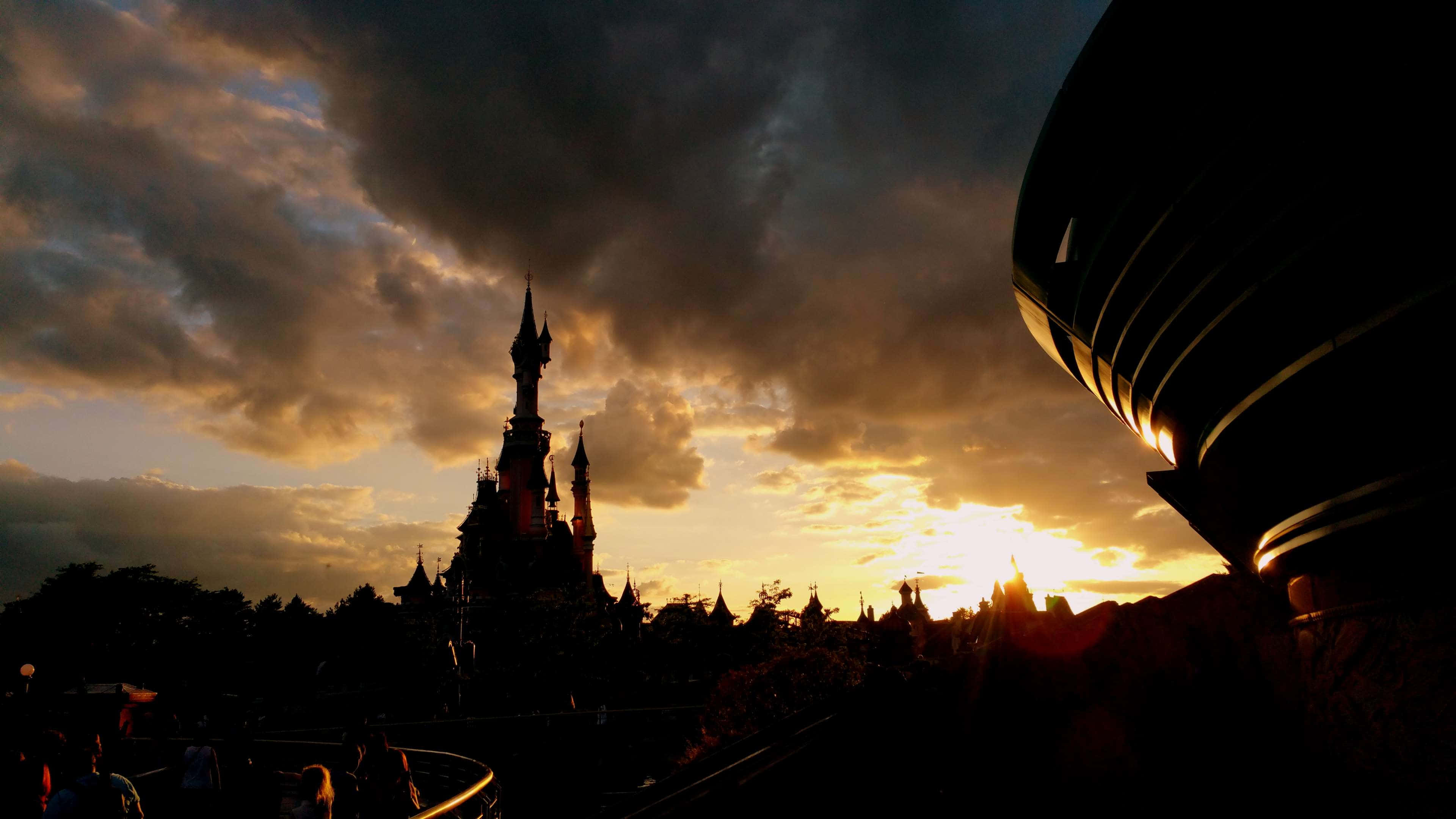 Disneyland 桌布 壁紙 — 一座雄偉的城堡在金色雲彩的戲劇性日落天空下形成剪影，前景有一座現代建築。